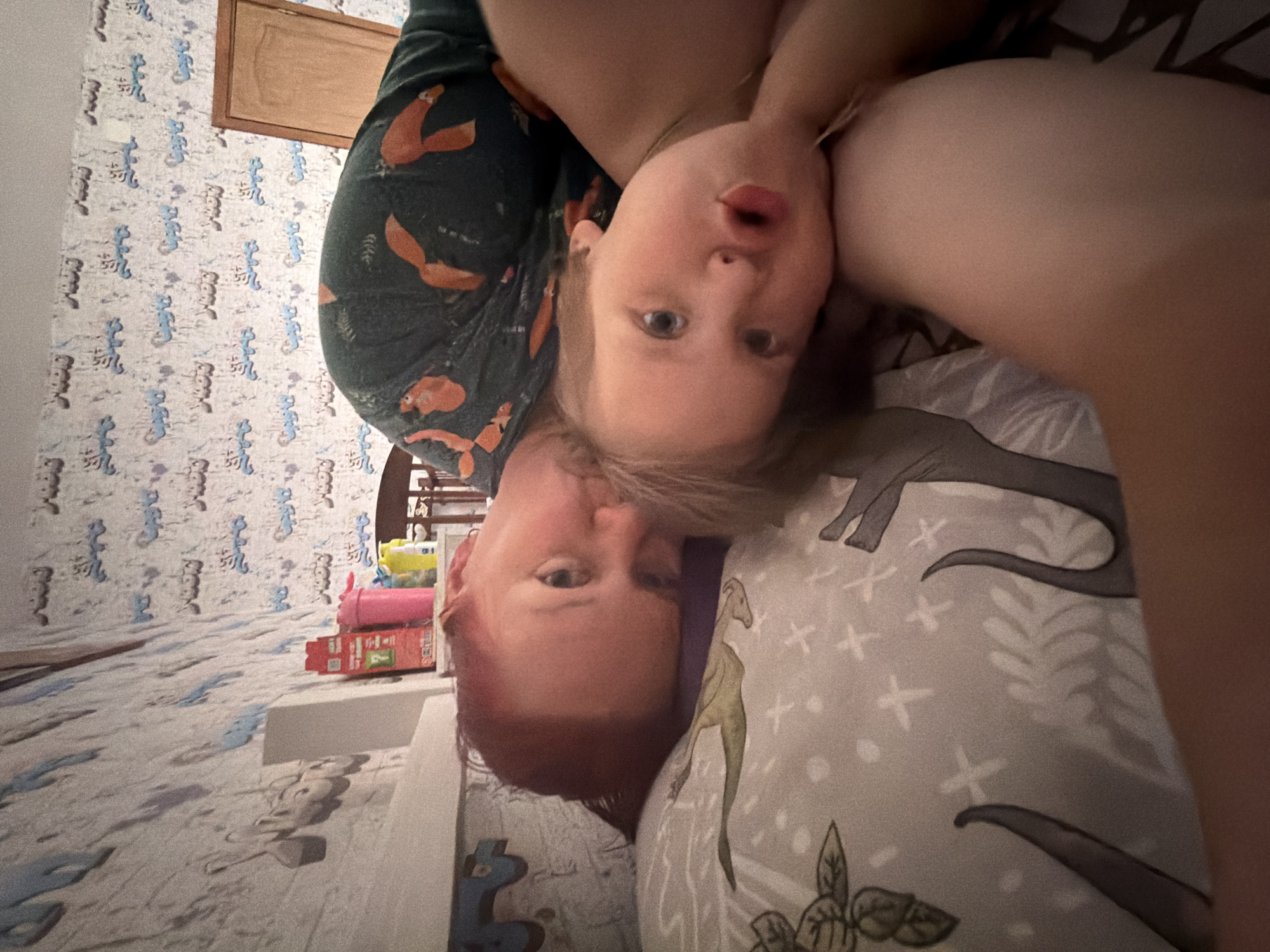 A selfie taken indoors in Chaikovsky, Russia shows two people: a fair-skinned child with blonde hair and blue eyes looking at the camera with an open mouth, and an adult with fair skin and red hair turned upside down in relation to the child. The child is wearing a dark green shirt with orange figures. They are positioned on a bed with a light grey dinosaur-patterned pillow. In the background, a wall is decorated with patterned wallpaper. A wooden cabinet and a shelf with various toys and objects, including pink and yellow items, are also visible.