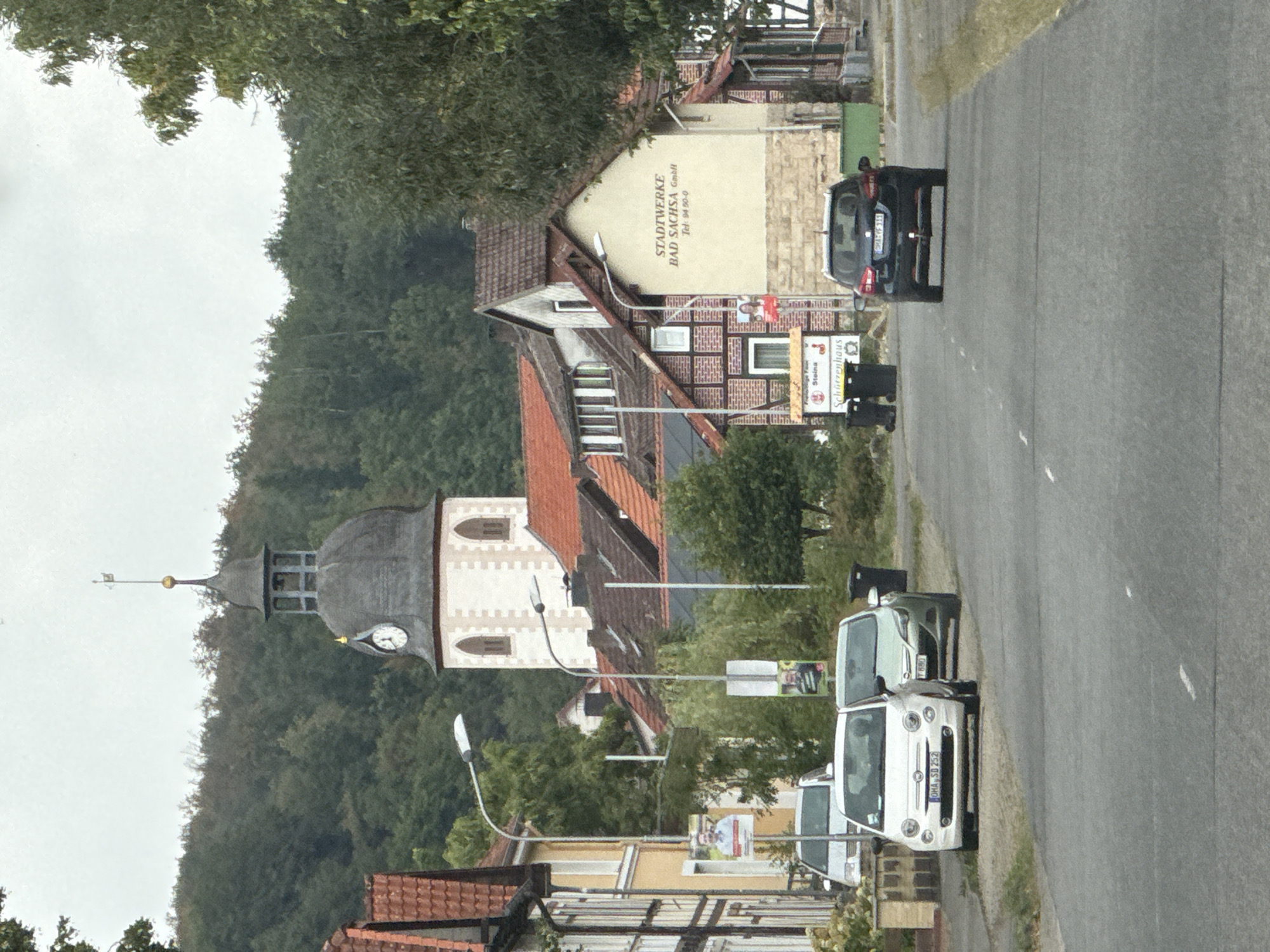 The image captures a street scene in Bad Sachsa, Germany, with a prominent church tower in the background. The tower has a dark, multi-tiered spire topped with a weather vane. A clock face is visible on the tower. In the foreground, there is a paved road with several parked cars. A white Fiat 500 is parked on the left, followed by a silver hatchback. Further down the road, a dark-colored SUV is driving away from the camera. Buildings line the street, some featuring traditional half-timbered architecture and others with more modern facades. One building has text that reads "STADTWWERKE BAD SACHSA GmbH". Campaign posters are visible on poles along the sidewalk. The environment appears to be a town or village setting, with trees and a slightly overcast sky.