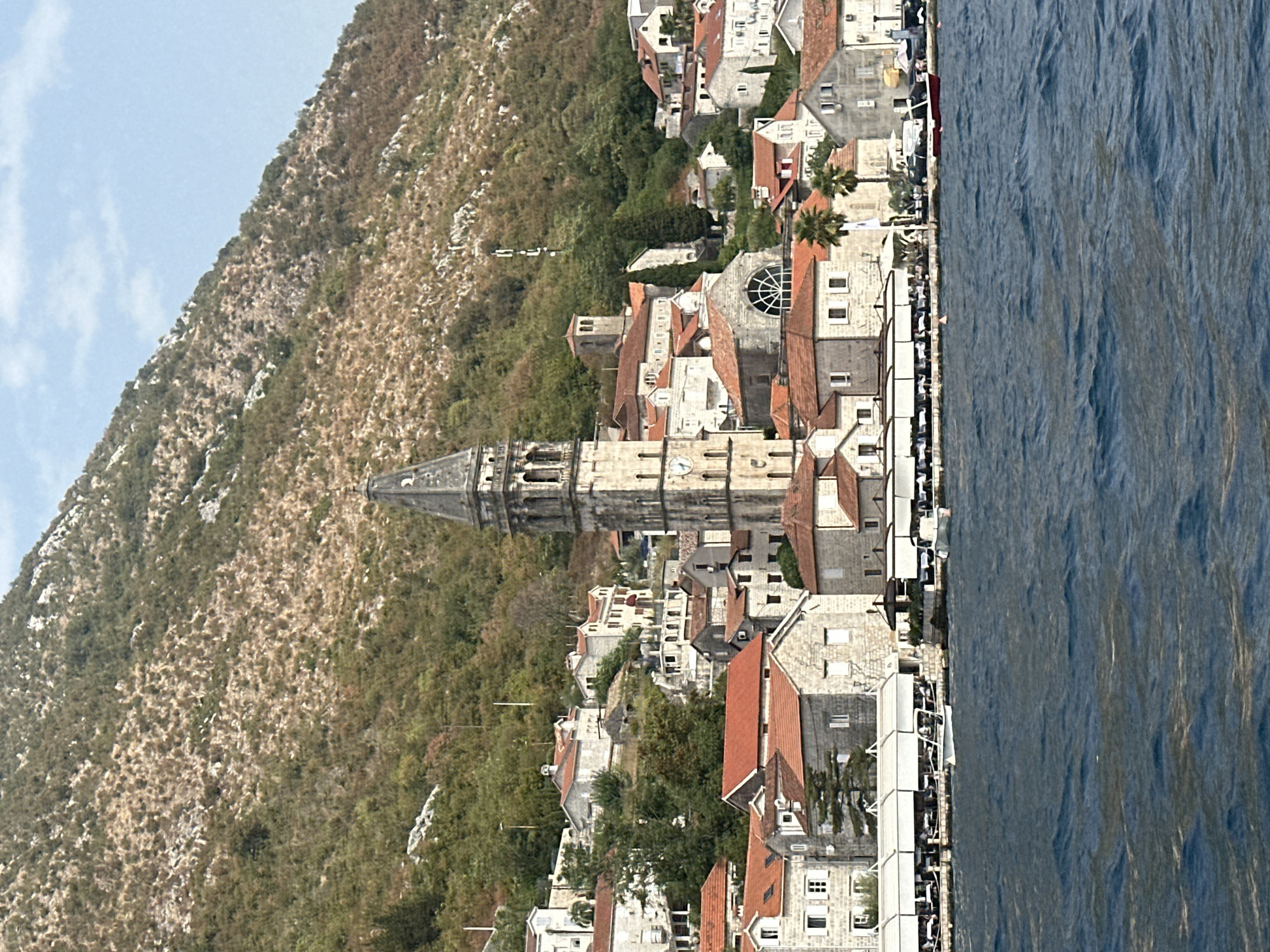 The image shows a view of the town of Perast, Montenegro, from the water. In the foreground, the deep blue water of the bay is choppy with whitecaps. Behind the water, a waterfront promenade is lined with buildings constructed of light-colored stone with red tile roofs. Many of these buildings appear to be restaurants, indicated by the awnings and outdoor seating arrangements. Several small boats are moored along the waterfront, including a white speed boat and a small red and white vessel.
Dominating the scene is a tall, ornate bell tower with a pointed spire, likely part of a church. The tower is made of weathered stone and features arched openings and a clock face. Further up the hillside behind the town, the slopes are covered in dense vegetation and rocky outcrops. The houses and buildings in Perast are clustered together, with some built on terraces on the hillside, creating a picturesque coastal town setting. The sky above is a pale blue with some wispy clouds. The overall impression is of a historic European coastal town on a sunny day.