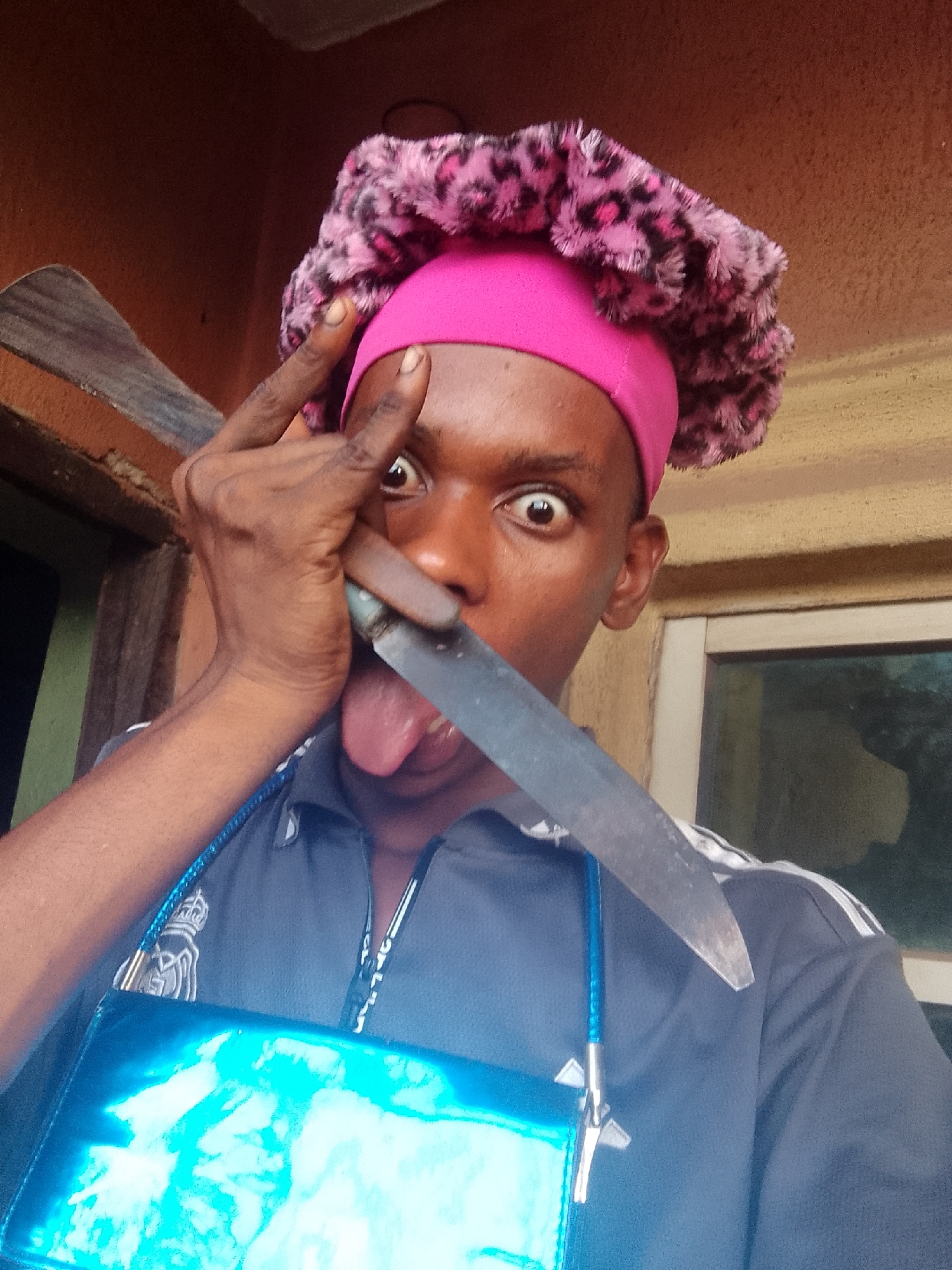 A man is holding a knife near his face. He is wearing a pink and leopard print bonnet and a blue Adidas track jacket. He has his tongue sticking out and his eyes are wide. There is a blue bag hanging from his shoulder. The location appears to be indoors, with a window visible in the background. The city and specific location are unknown.