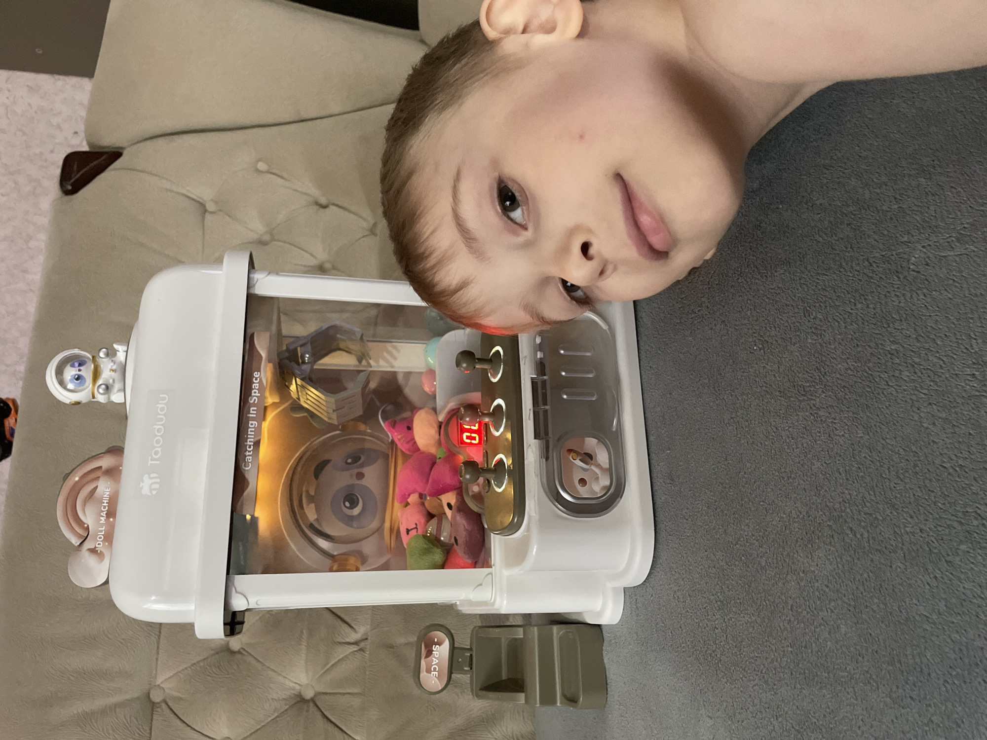 A fair-skinned child with short, light brown hair is lying on a gray surface and looking into the camera. The child is positioned at the right side of the frame. To the left of the child is a white toy claw machine, which is filled with plush toys. The claw machine has the words "Taodudu" and "Catching in Space" printed on it. A digital display on the machine shows the number "10." The scene is indoors, with a tan cushioned surface visible in the upper left of the frame. The location or presence of Baranovka, Russia cannot be verified from the image alone.