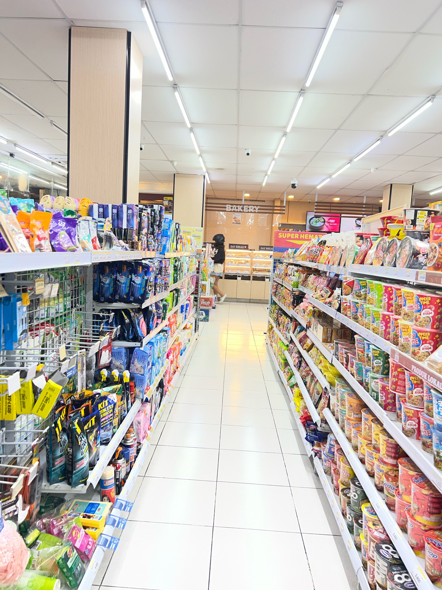 The image shows the interior of a well-lit convenience store. The aisle is lined with shelves stocked with various packaged goods, primarily food items like instant noodles and snacks, as well as cleaning supplies. Fluorescent lights are mounted on the ceiling, illuminating the white tiled floor and the brightly colored products. A person, a woman with dark hair wearing a black top and a light-colored skirt, is walking down the right aisle, viewed from behind. She is holding a shopping basket. Another person, possibly an employee, is visible in the background near a "BAKERY" sign and display cases. The overall scene suggests a typical retail environment in Denpasar, Indonesia, indicated by the text on the signage like "SUPER HEMAT" (Super Save) and "PRODUK LOKAL" (Local Products).