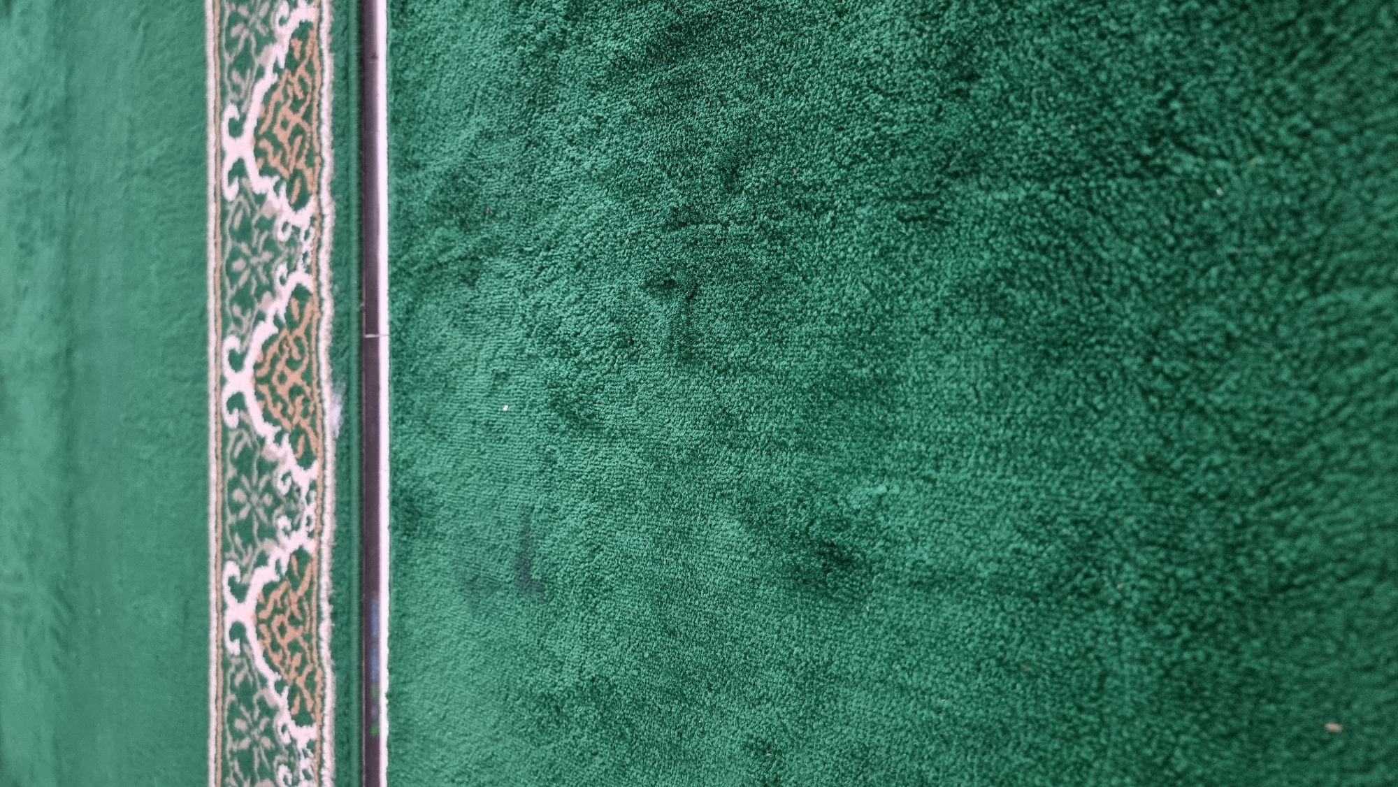 The image displays a close-up view of a green carpet, likely a prayer rug, with a decorative border. The border features intricate patterns in white and gold. The carpet appears to be made of a soft, looped material. There are a few dark marks or stains visible on the green carpet. The perspective is from a low angle, looking across the surface of the carpet. The environment is indoor, and the lighting is even, suggesting ambient light rather than direct sunlight. The specific location in Meunasahlhok, Indonesia is not discernible from the image itself. There are no people, actions, or interactions depicted in the scene.