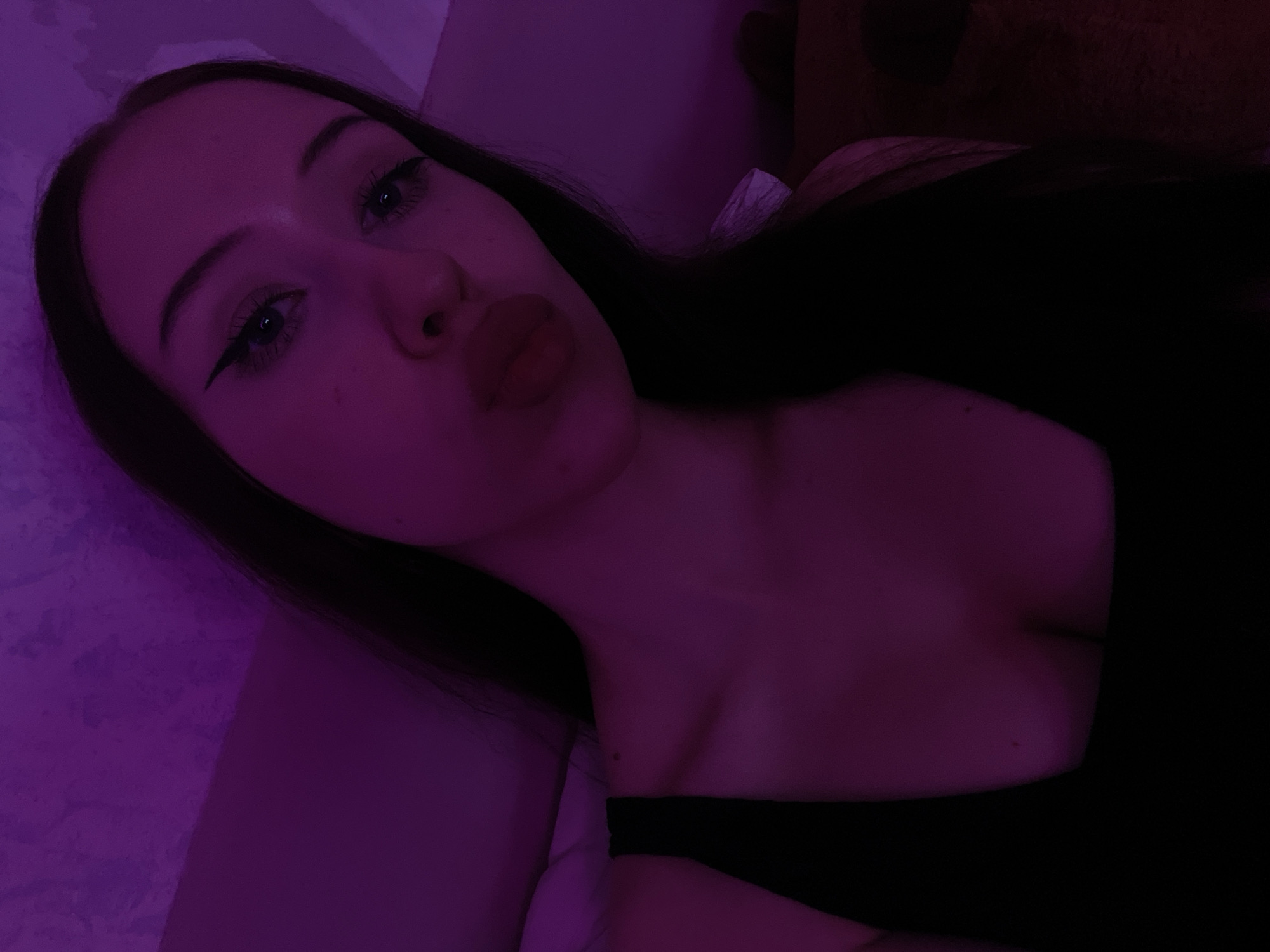 A young woman with long dark hair and winged eyeliner poses for a selfie in a dimly lit room. She is wearing a black top that reveals her collarbone and upper chest. The lighting in the room is predominantly purple, casting a violet hue over the scene. The background appears to be a textured wall and possibly a bed or soft surface. The woman's lips are slightly pursed as if blowing a kiss. The image was taken indoors.