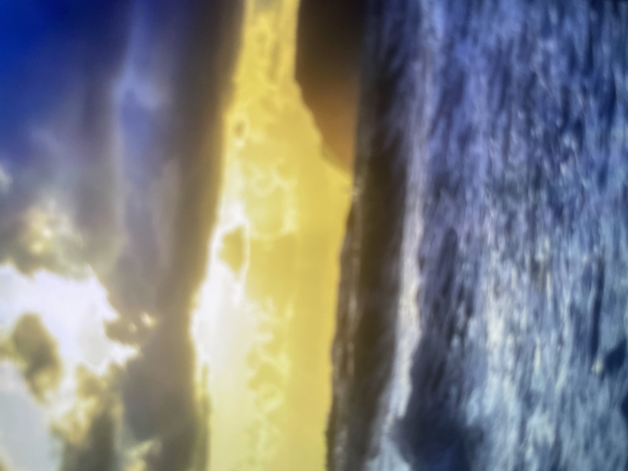 The image depicts a blurry seascape at sunset. The sky is a dramatic mix of dark clouds tinged with yellow and blue, illuminated by the setting sun which appears as a bright, hazy orb behind a silhouetted landmass. The ocean in the foreground is rough, with visible waves and whitecaps, rendered in shades of dark blue and grey. The overall impression is one of a stormy, yet beautiful, natural scene. There are no people or man-made structures visible. The location is not Khanty-Mansiysk, Russia.