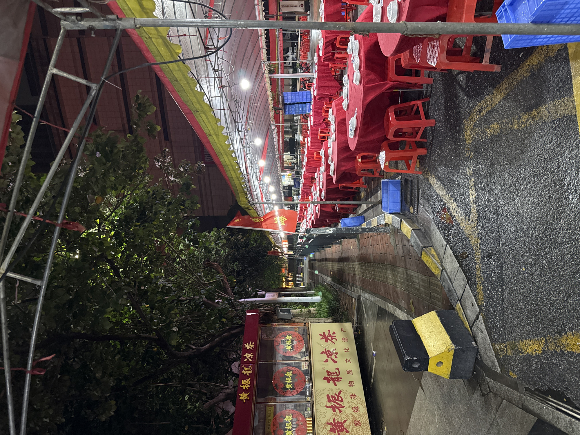 The image depicts an outdoor dining setup at night in Haizhu District, China. A large tented area with red tablecloths covers multiple round tables, each set with white bowls. Red plastic stools surround the tables. The ground is wet, suggesting recent rain, and yellow markings are visible on the asphalt. To the left, a small stand displays signage with Chinese characters and circular emblems. A tree with dense foliage is situated behind the stand. A black and yellow traffic barrier is positioned on the sidewalk. The scene is illuminated by overhead lights within the tent.