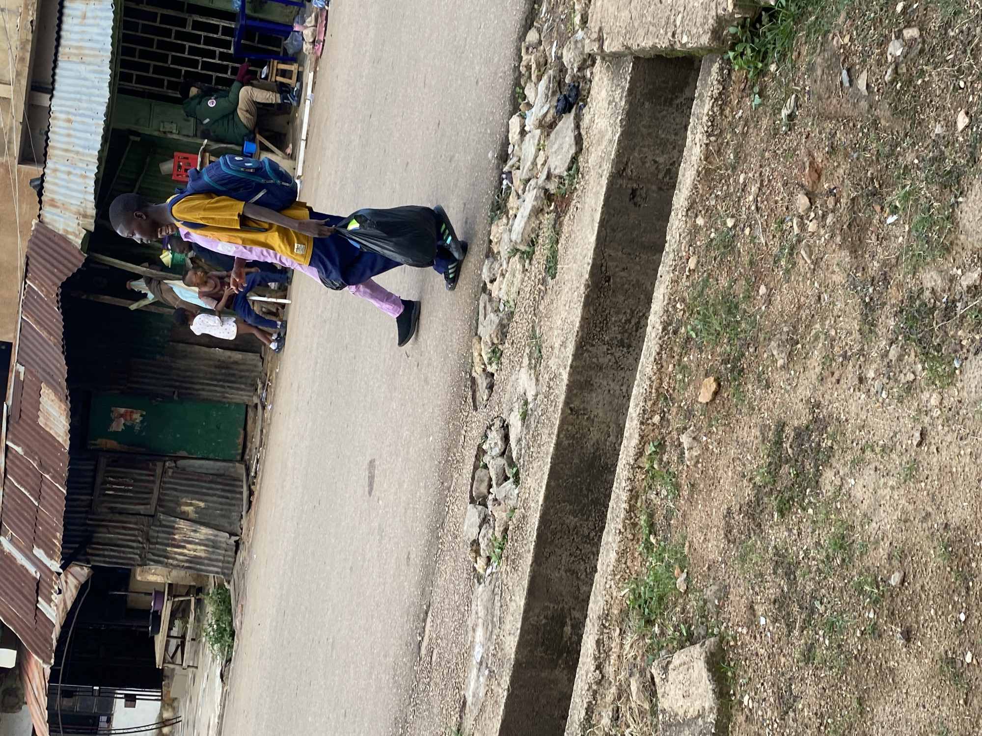 The image depicts a street scene in Jos, Nigeria, featuring a group of people near a roadside structure. The structure, made of corrugated metal and wood, appears to be a shop or market stall with a rusted tin roof. A young boy in a yellow and pink shirt, carrying a blue backpack and a black plastic bag, walks across the foreground, his attention directed towards the left. Several other children are visible in the background, one wearing a white shirt and another in a brown shirt. A man in a green jacket sits on a stool to the right, and other individuals are present further back. The street is paved, and a concrete drainage ditch runs along the side, filled with rocks and debris. The ground is a mix of dirt, gravel, and sparse vegetation.