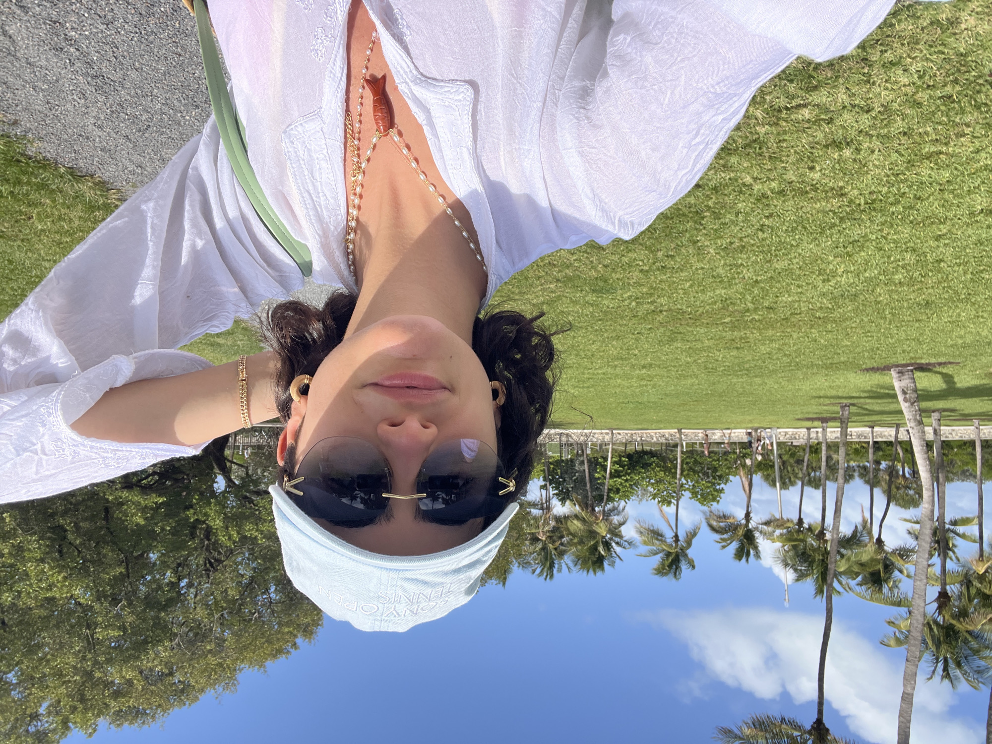 The image is an upside-down selfie taken outdoors. The subject is wearing a white top, sunglasses, and a light blue "Sony Open Tennis" hat. A beaded necklace with an orange pendant is visible. The background features green grass, palm trees, a clear blue sky with scattered clouds, and what appears to be a building or structure with people in the distance. The location is likely Miami Beach, United States, as indicated by the "Sony Open Tennis" hat.