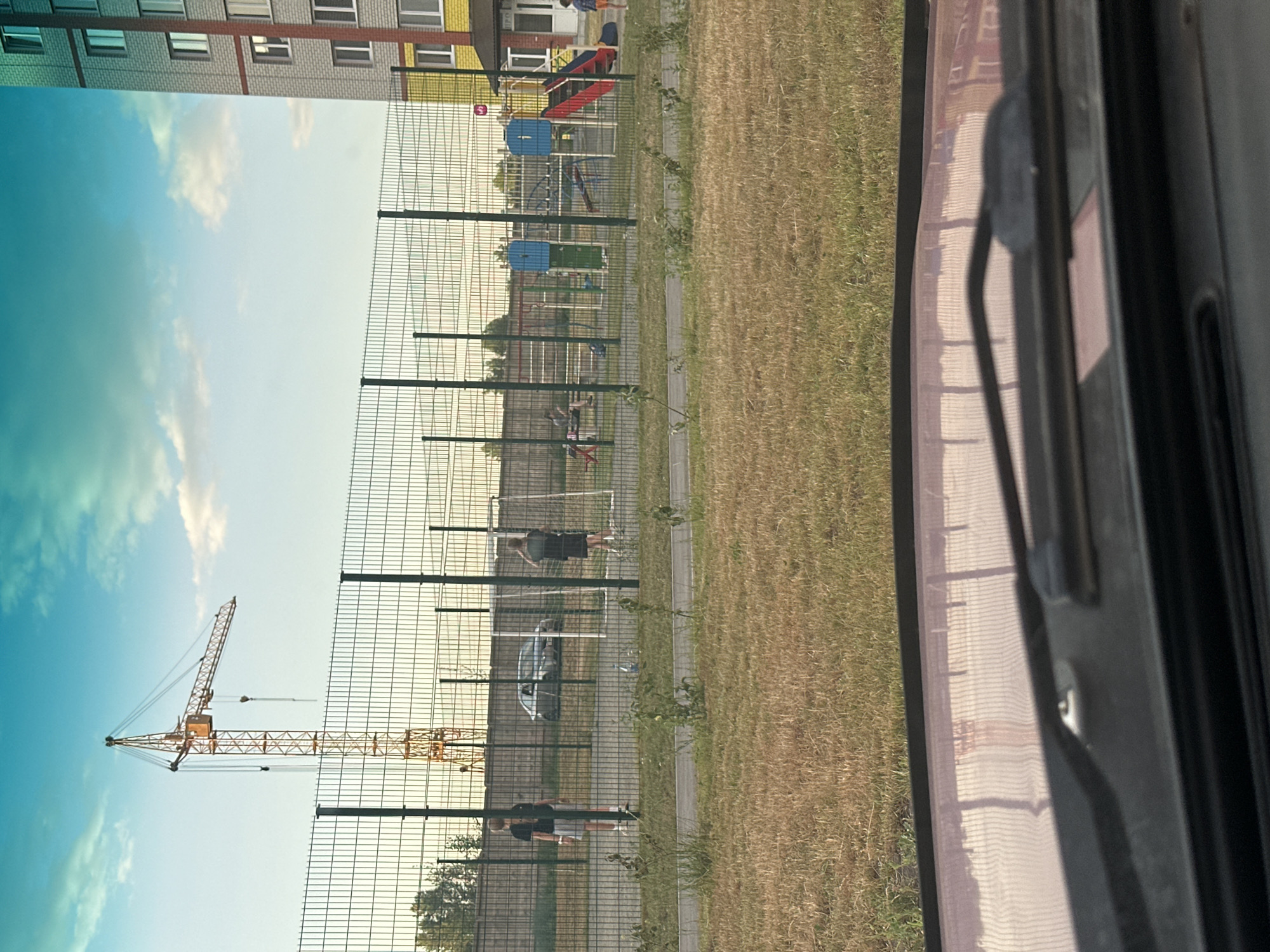 The image shows a construction crane against a bright sky with clouds, located behind a chain-link fence. To the right of the crane, a multi-story building with red and white accents is visible. Inside the fenced area, a playground with slides and climbing structures is present. Two individuals, one in a dark t-shirt and shorts and another in a dark t-shirt and shorts, appear to be playing or exercising in the yard. A dark-colored car is parked in the background. The foreground of the image shows the reflection of the fence on a car's windshield, indicating the photo was taken from inside a vehicle. The environment appears to be an outdoor urban setting in Novoaltaysk, Russia, given the presence of residential buildings and a construction site.