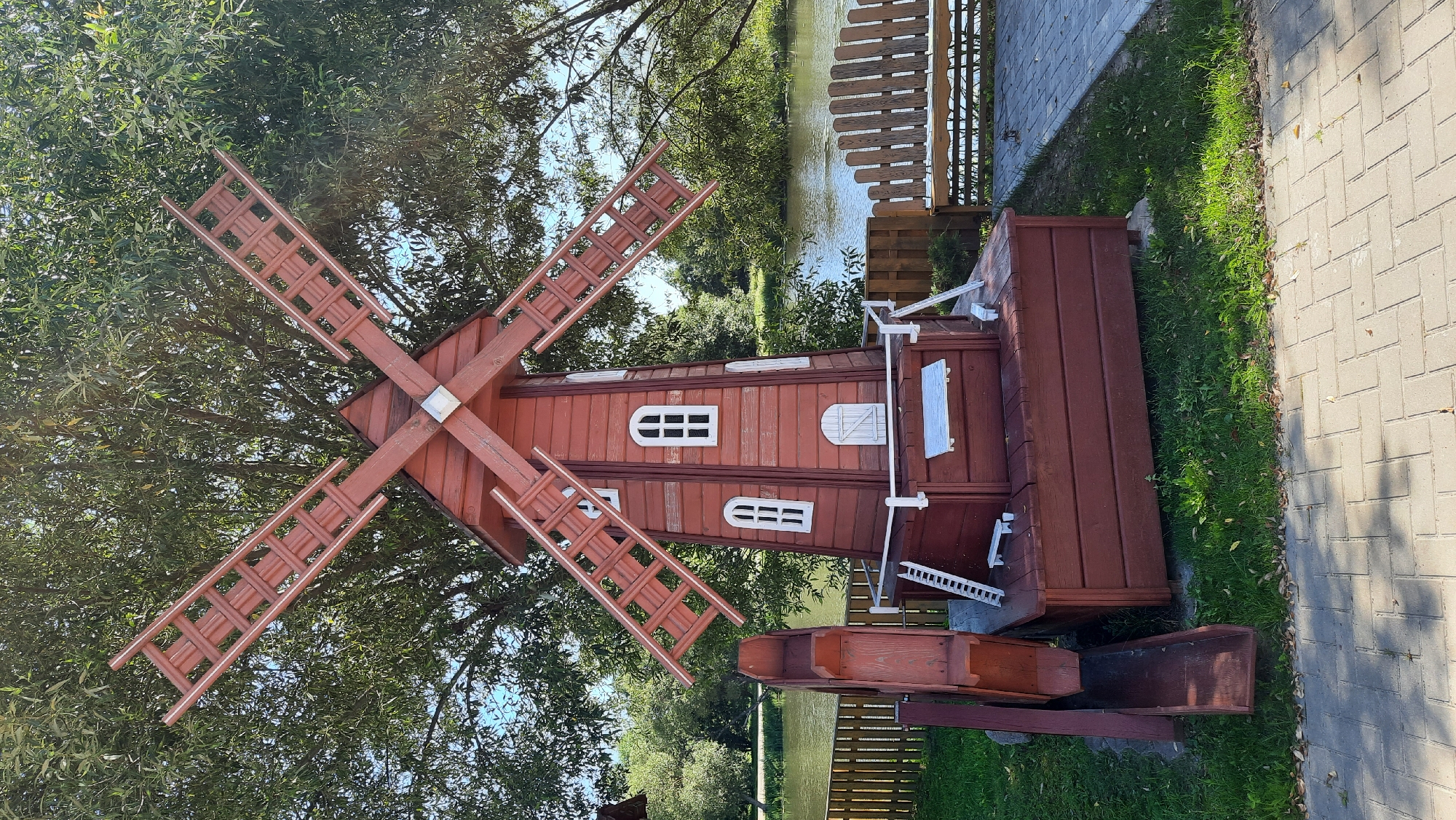 An eye-level, medium shot captures a decorative red wooden windmill situated outdoors on a sunny day. The windmill, a prominent feature, stands against a backdrop of lush green trees and a body of water, possibly a river or lake. To the left of the windmill is a wooden water wheel with a chute. A wooden fence runs along the grass behind the windmill, and beyond that, a wooden bench and a paved walkway are visible. The ground in front of the windmill is paved with light-colored interlocking bricks, and the area directly around the windmill is covered in green grass. The sky is a clear blue.