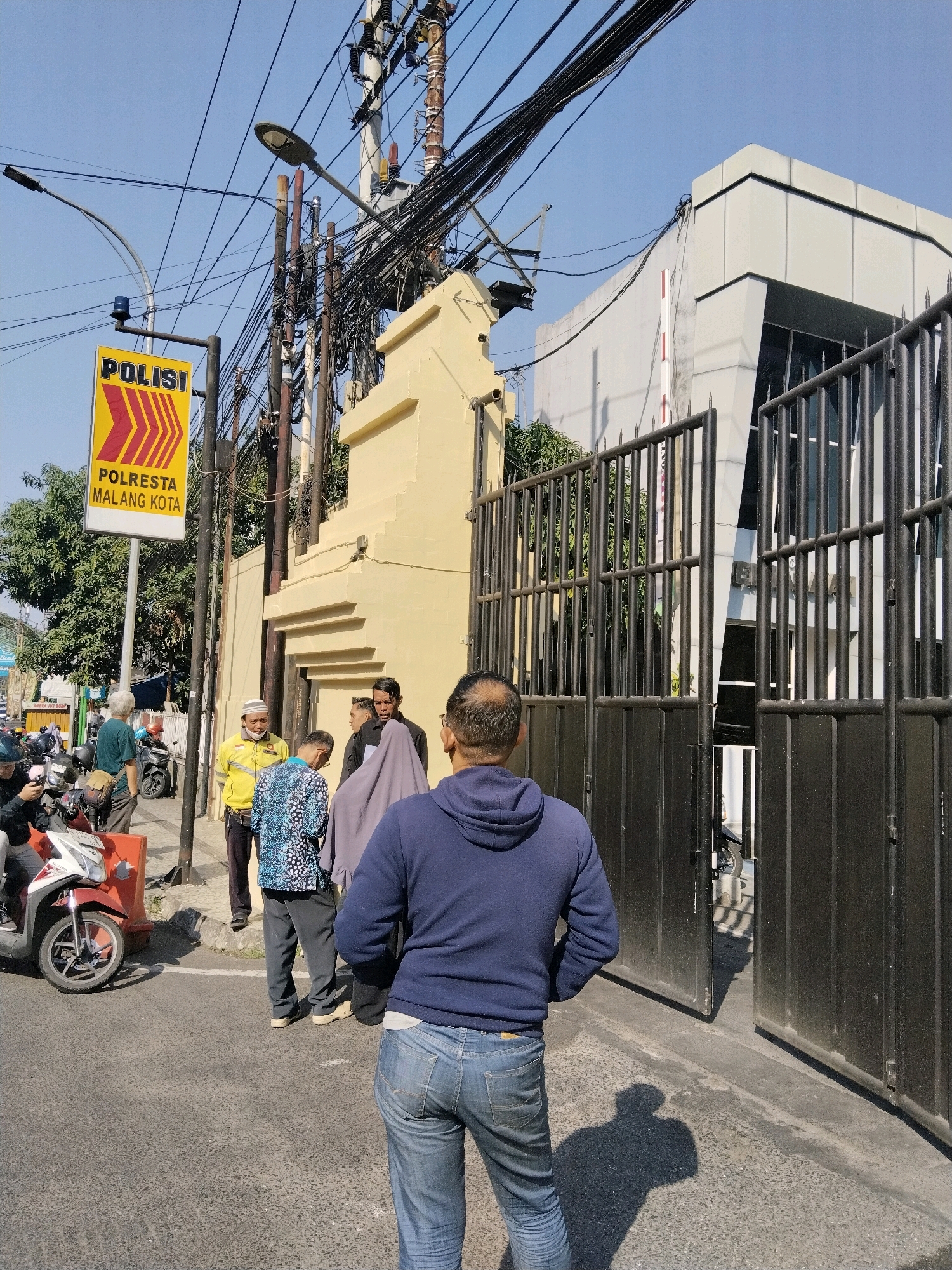 The image depicts the entrance of the Polresta Malang Kota police station in Malang, Indonesia. A yellow sign with red arrows and the word "POLISI" (Police) is prominently displayed on the left. Below it, the text "POLRESTA MALANG KOTA" indicates the specific police station. A group of men are gathered near the entrance. One man in the foreground, facing away from the camera, wears a blue hoodie and jeans. To his left, a man in a blue and white patterned shirt and dark pants stands near a man in a yellow jacket and white cap. Another man in a dark jacket and a person in a grey headscarf are also present. The entrance is secured by a tall black metal gate, partially open. The building behind the gate is modern with grey panels. Overhead, a complex network of electrical wires crisscrosses against a clear blue sky. Motorcycles are visible parked on the street and near the entrance, suggesting a busy urban environment. The overall scene captures a moment outside a public service building in a city.