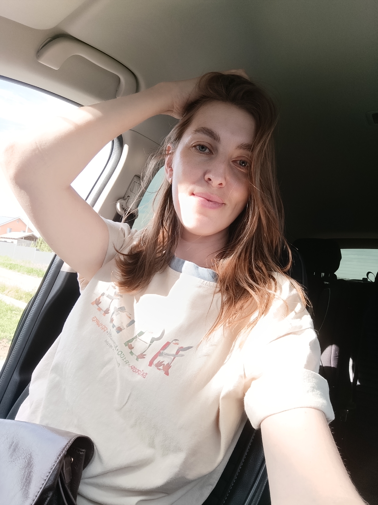 A medium shot shows a woman seated in the driver's seat of a car. The woman has fair skin and brown hair. Her left hand is touching the top of her head. She wears a light-colored t-shirt with a graphic design of stylized figures in red, green, and black. The car's interior is predominantly black and gray. The window behind her reveals a street with green grass and a few houses in the background. The image suggests the location could be Chaikovsky, Russia.