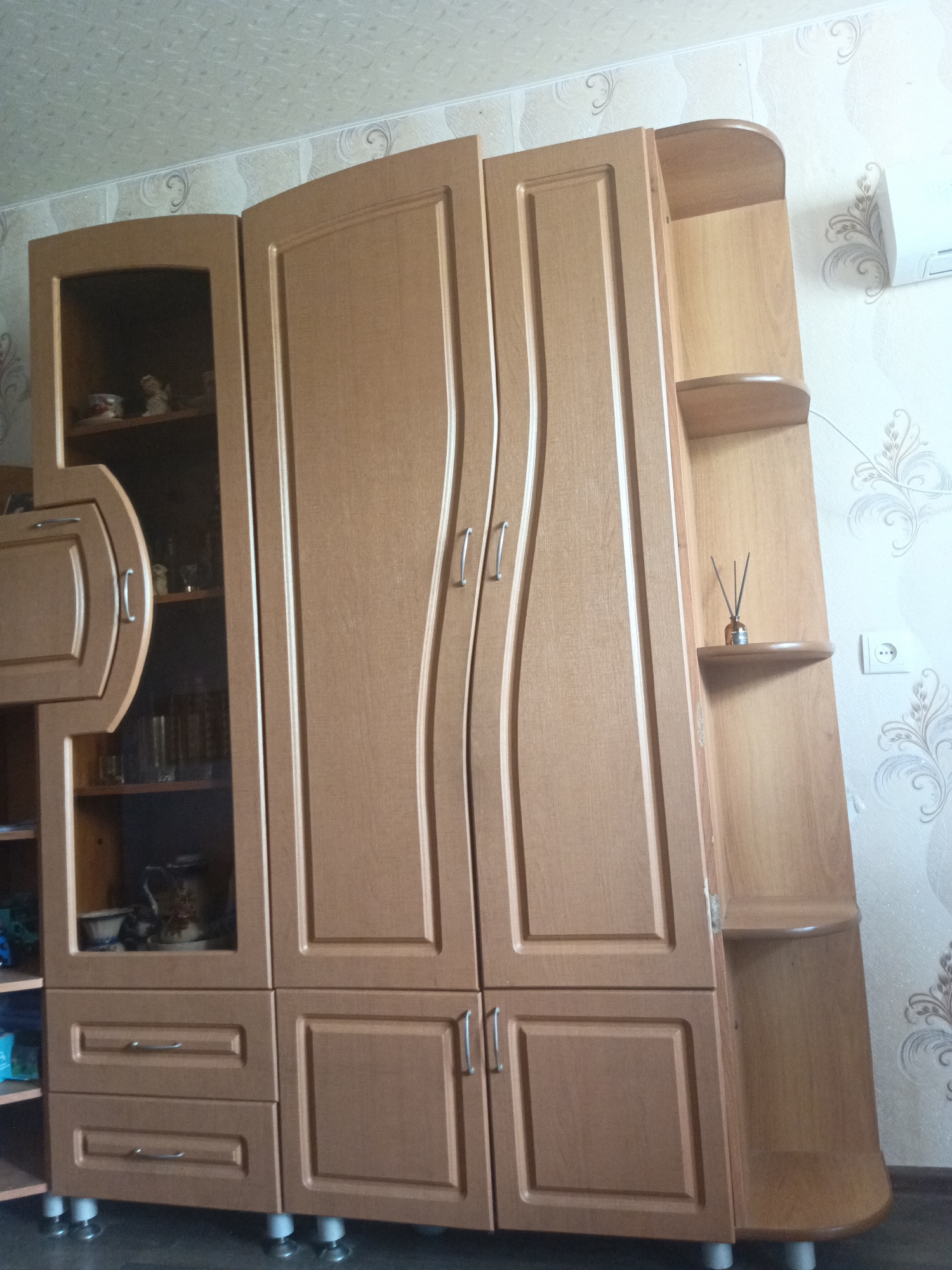 The image shows a light brown wooden wardrobe in a room. The wardrobe has multiple doors and drawers, and a glass-fronted section on the left. On the right side of the wardrobe, there are open shelves. On one of these shelves, a small glass bottle with diffuser reeds is visible. A white air conditioning unit is mounted on the wall above the right side of the wardrobe. The walls are covered with light-colored wallpaper with a floral pattern. The floor is dark and appears to be tiled or linoleum. The wardrobe stands on small, round white legs. The lighting in the room appears to be from overhead. The scene is consistent with a domestic interior.