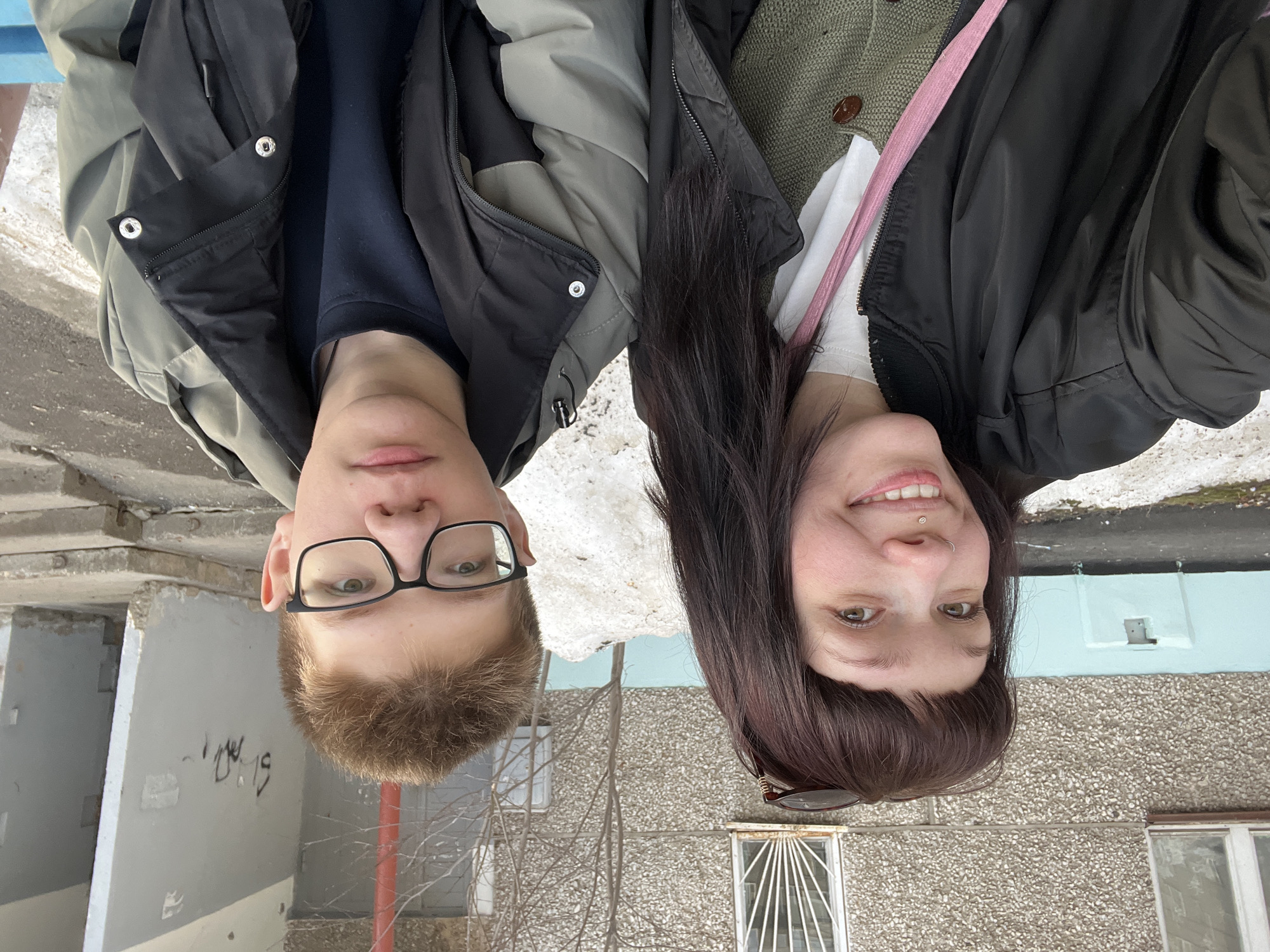 An outdoor scene in Izhevsk, Russia, depicts two individuals positioned for a close-up photograph, likely a selfie, on a cold-weather day.
**People:**
1. **Female Subject (Left):** A young adult female with dark brown, shoulder-length hair and bangs, light skin, and brown eyes, is looking towards the camera with a smile. She has a silver stud in her left nostril and a gemstone stud in her philtrum. Sunglasses are resting on top of her head. She wears a black bomber jacket over a white crew-neck top and a green knitted cardigan with a visible brown button. A pink fabric strap, possibly from a bag, crosses her chest.
2. **Male Subject (Right):** A young adult male with light brown, short hair, light skin, and a neutral expression, is looking towards the camera. He wears black-rimmed glasses. His attire consists of a multi-toned jacket, primarily olive green and black, over a dark blue or black long-sleeved shirt.
**Actions/Interactions:** The individuals are seated side-by-side, occupying the central and lower-middle portions of the frame, facing the camera. Their close proximity suggests an interpersonal relationship.
**Environment and Objects:** The background features the facade of a multi-story building constructed with a textured, pebbled concrete finish. Visible elements include a window with a white frame and white metal security bars on the left, and another window on the right. A lower section of the building's wall is painted a light teal or blue color, with a small rectangular vent or access panel. Dirty snow piles are present on the ground behind the subjects and along the building's base, interspersed with patches of bare earth or pavement. A leafless deciduous tree or bush stands behind the subjects. To the far right, leaning grey concrete slabs, possibly construction remnants or barriers, are visible, exhibiting some dark graffiti or markings. A reddish vertical pipe or pole is partially visible on the far right edge. A thin strip of a blue surface, likely a bench, is visible at the bottom right corner of the frame, underneath the male subject. The lighting is diffused, indicating an overcast sky or indirect sunlight.
