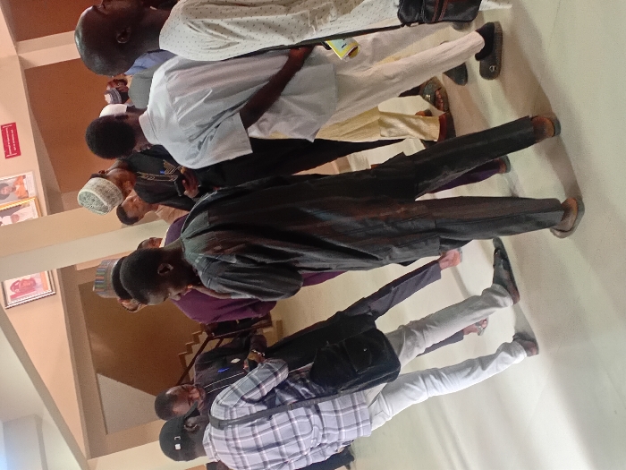The image displays an indoor scene in the city of Kwazailewa, Nigeria, populated by approximately ten to fifteen men. The setting is a brightly lit hall or lobby with a smooth, light-colored floor and neutral-toned walls. In the upper background, two framed portraits and a small red rectangular sign are mounted on a wall. A dark-colored staircase with a visible handrail ascends on the left side of the background.
The individuals are primarily young to middle-aged adult males, dressed in a combination of traditional Nigerian garments, such as kaftans and agbadas, and casual attire including shirts and trousers. Several men wear traditional caps.
In the immediate foreground on the left, a man wearing a light plaid shirt, light trousers, and a dark cap carries a black shoulder bag and is focused on a smartphone. Adjacent to him, another man, seen from behind, wears a dark, possibly striped, traditional two-piece outfit and open-toe sandals. To the right of the foreground, a man in a dark traditional outfit and a white/grey patterned cap is looking down at a smartphone. Further back, another man is seen from behind, wearing a light blue/grey short-sleeved shirt and light trousers. Most individuals are standing, with several observed examining their smartphones. No direct physical interactions or explicit conversations are discernible.