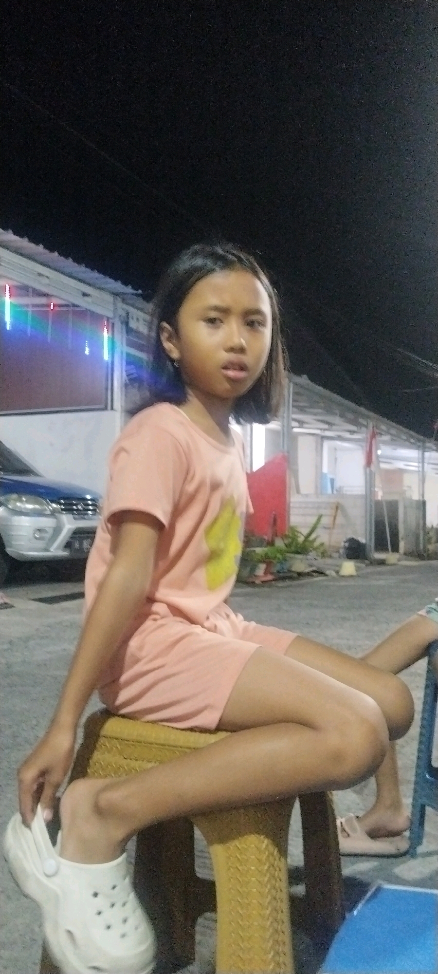 The image shows a young girl sitting on a yellow woven stool in an outdoor setting at night. She is wearing a peach-colored t-shirt with a yellow graphic and matching peach-colored shorts. The girl has dark hair styled in a bob and is looking towards the camera with a neutral expression. Her right foot is resting on her left shoe, which is a white clog. In the background, there is a building with white walls and a corrugated metal roof. Colorful lights are strung across the facade of the building. To the left of the girl, part of a blue car is visible. The ground appears to be paved. The overall scene suggests a casual evening gathering or a commercial area illuminated by decorative lights. The presence of the building, car, and outdoor seating implies a semi-urban or commercial environment, potentially a street food stall, a small shop, or a residential area with an outdoor seating arrangement in Semarang, Indonesia.