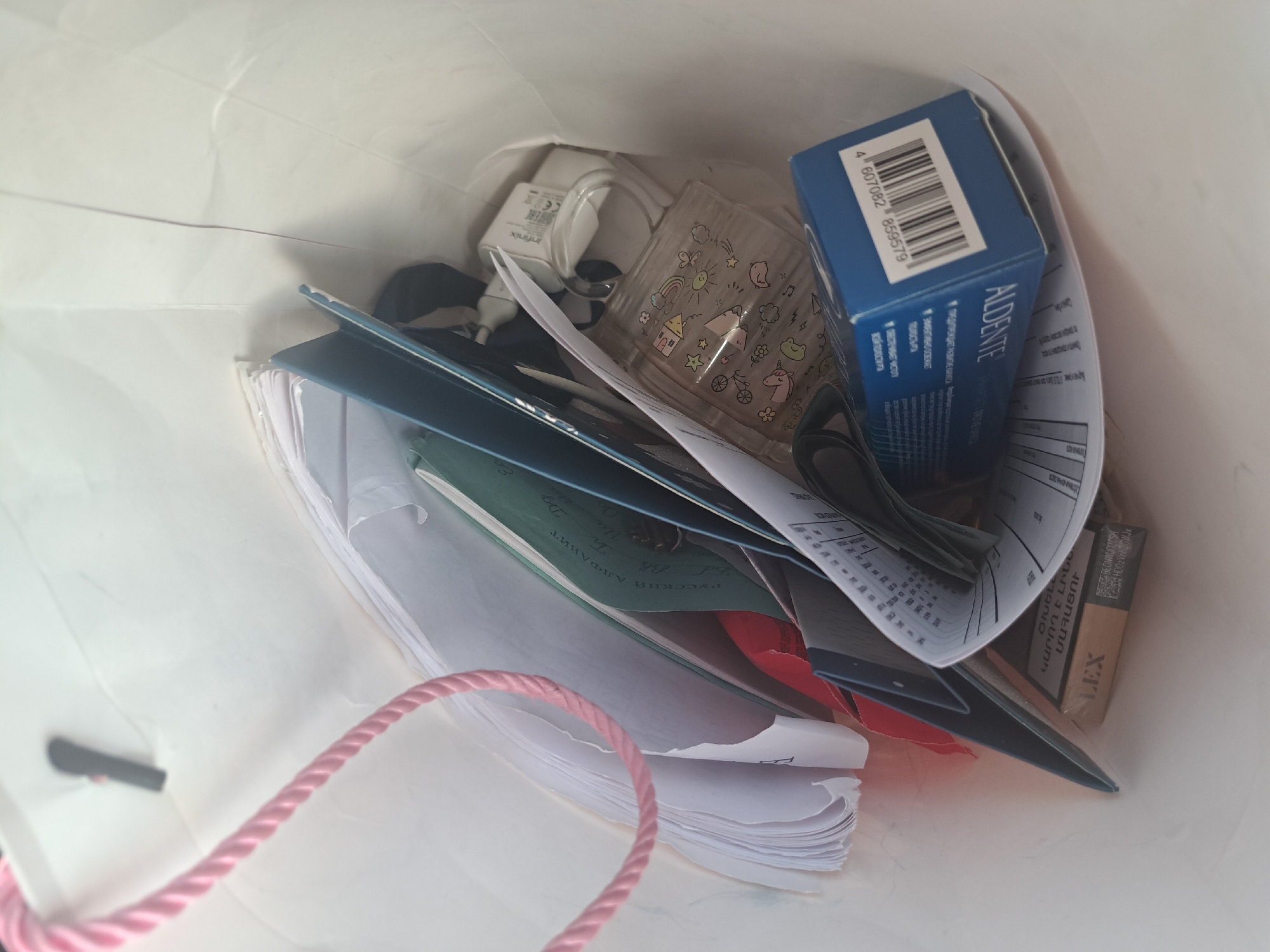 This is an eye-level, indoor, medium shot of a white paper shopping bag filled with various items. The bag's left side has a pink braided rope handle. Inside the bag, from left to right, are several stacks of papers, some appearing to be documents or notebooks. Further in, there is a dark blue folder or binder. Next to it, a white USB wall charger with the brand "Infinix" is visible. To the right of the charger is a clear, rectangular plastic cup adorned with colorful cartoon drawings of houses, flowers, stars, and unicorns. Beneath the cup and charger, a small, clear glass bottle with a white lid is partially obscured. In the lower right of the bag is a blue cardboard box with the text "AL'DENTE" and a barcode. In the lower left, a pack of cigarettes with the brand "LEX" is visible. The background is the white interior of the shopping bag. The scene appears to be set in Leningradskaya, Russia, given the Armenian script on some of the documents, which suggests a connection to the Armenian community or businesses in the region, and the presence of the USB charger which could be used in that city.