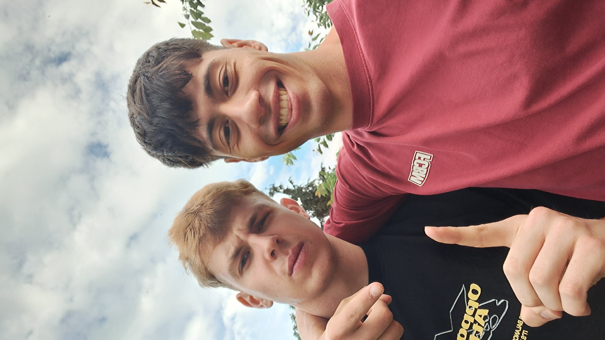 Two men are posing for a selfie outdoors. The man in the upper portion of the frame is smiling broadly, with dark hair and wearing a red t-shirt. The man below has light brown hair and a more serious expression, wearing a black t-shirt and holding one of his hands near his throat. The background shows cloudy skies and some greenery, likely trees. Without more information, it is not possible to place the location in Burgas, Bulgaria.