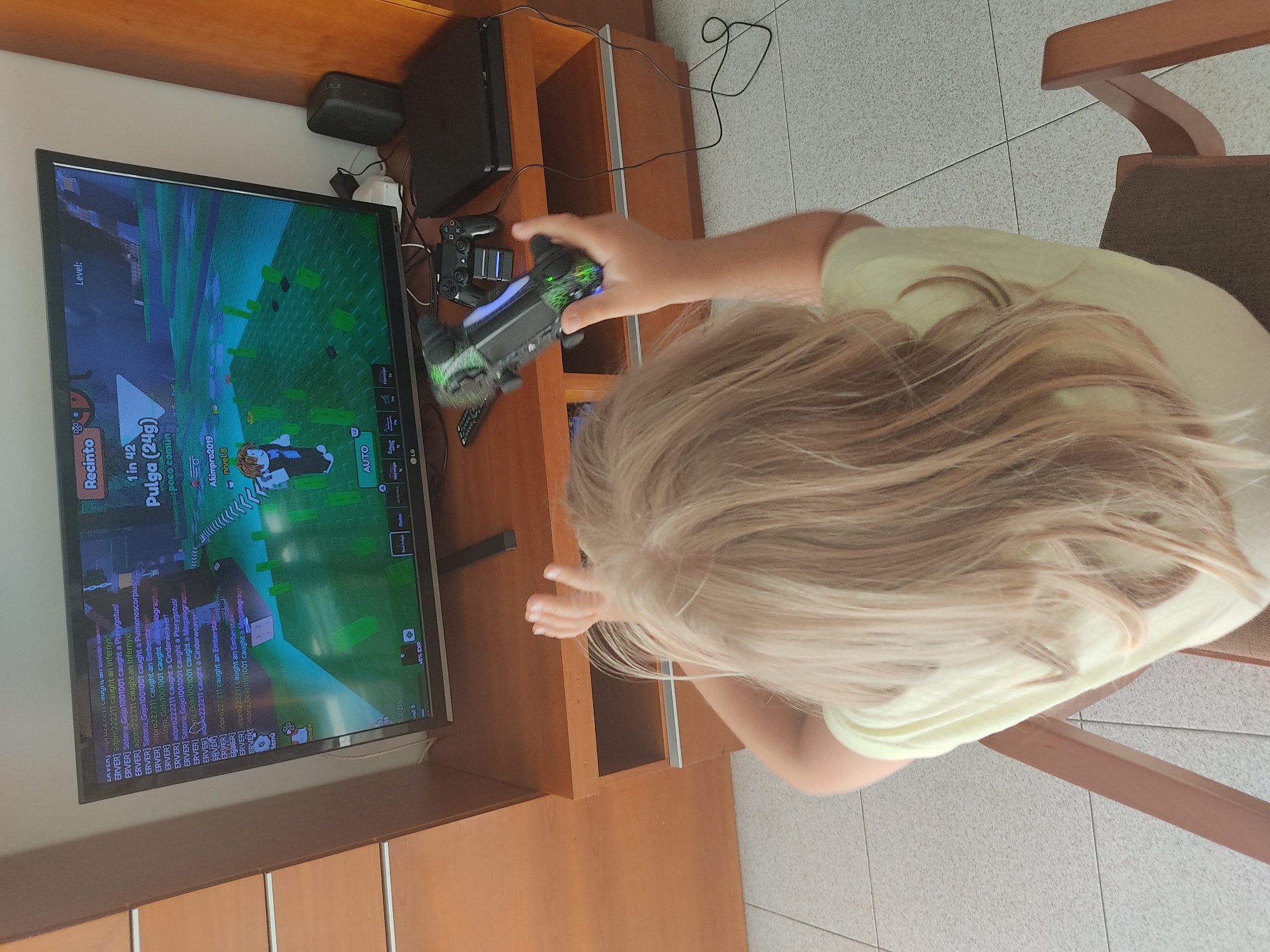 In Blanes, Spain, a young child with long, blonde hair is seated on a chair, facing away from the camera, engrossed in playing a video game on a television. The television displays a game with a chat log on the left side, detailing in-game actions and player names like "ERVER" and "adpro222211". The game itself appears to be a colorful, blocky environment with figures that resemble avatars. A PlayStation 4 console and two controllers are visible on the shelf beneath the television, suggesting the gaming setup. One controller, a black DualShock 4 with a green light strip, is held by the child. The setting appears to be a living room or entertainment area, with wooden furniture and tiled flooring.