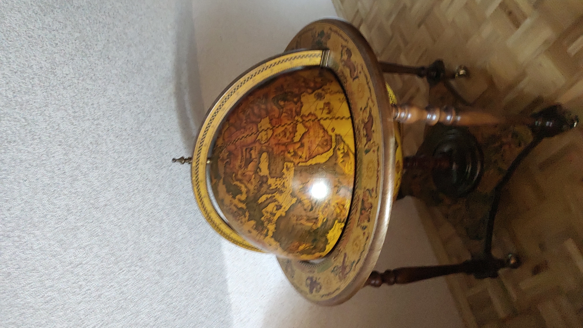 An antique terrestrial globe is displayed on a wooden stand with decorative carvings and wheels. The globe itself is detailed with a map of the world, featuring intricate lines representing landmasses and oceans. The stand is made of dark wood and has four legs that lead to a circular base. The environment appears to be indoors, with a textured white wall visible in the background and a parquet floor beneath the stand. There is no indication of this scene being located in the city of Allendorf, Germany.