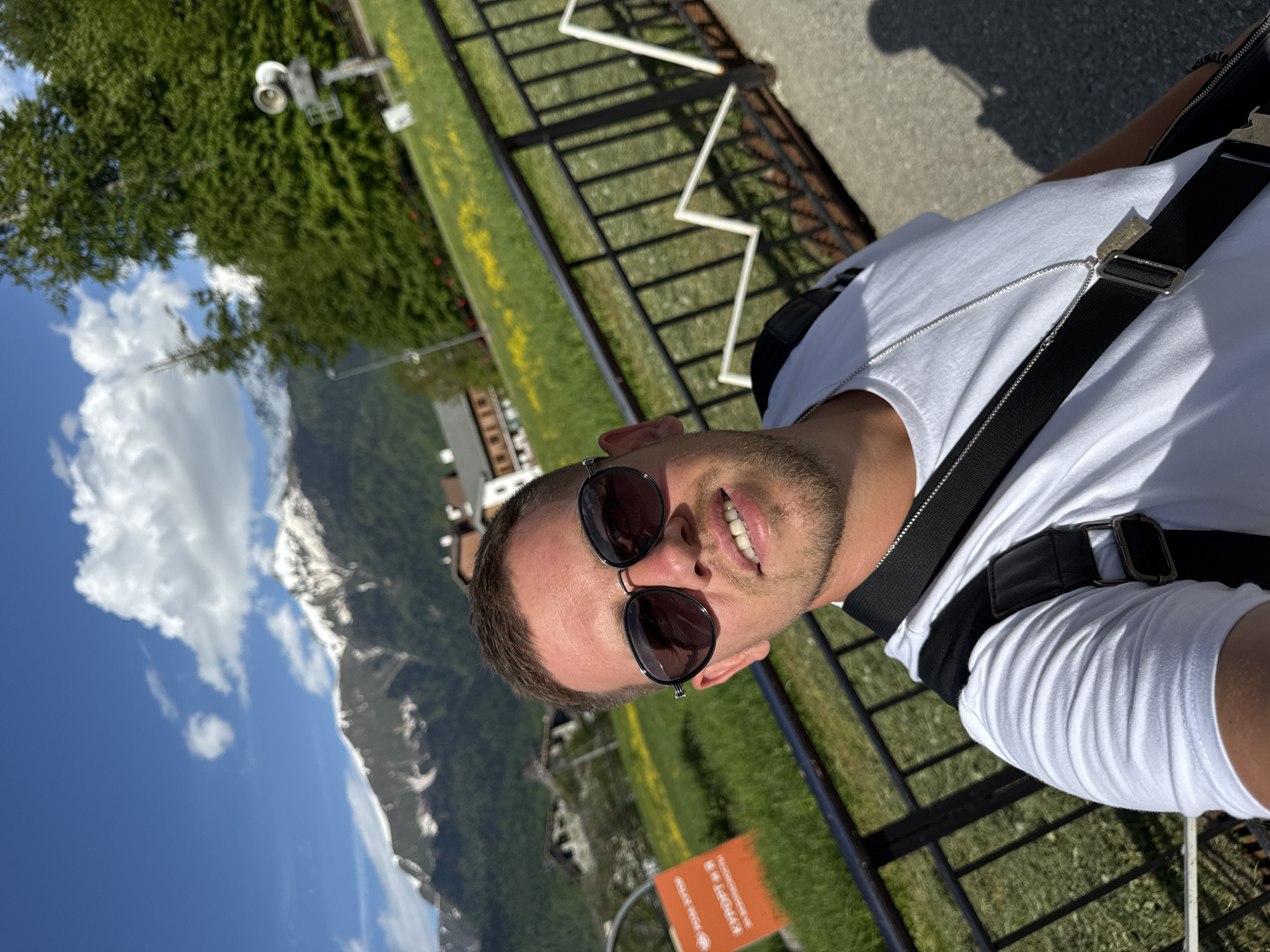 The image displays a man posing for a photo outdoors in a scenic mountain setting in Estosadok, Russia. The man is positioned in the foreground, looking directly at the camera with a slight smile, revealing his teeth. He is wearing dark sunglasses, a white t-shirt, and a black shoulder bag with a prominent strap across his chest. A thin silver chain with a small pendant is visible around his neck.
Behind the man, a black metal fence delineates a paved pathway from a lush green lawn interspersed with small yellow flowers. Further into the background, several buildings, consistent with a resort or village architecture, are visible. The most striking feature of the background is the majestic range of snow-capped mountains under a clear blue sky dotted with fluffy white clouds, indicating bright and sunny weather during the daytime. A small weather station or camera mounted on a pole is also visible on the left side, partially obscured by trees.
In the lower right corner, a portion of an orange sign is visible. The legible text in Cyrillic script reads "KVAPOPT M.S", which likely translates to "Resort M.S", fitting the context of a mountain resort area. The scene conveys a pleasant outdoor experience in a picturesque mountainous landscape.