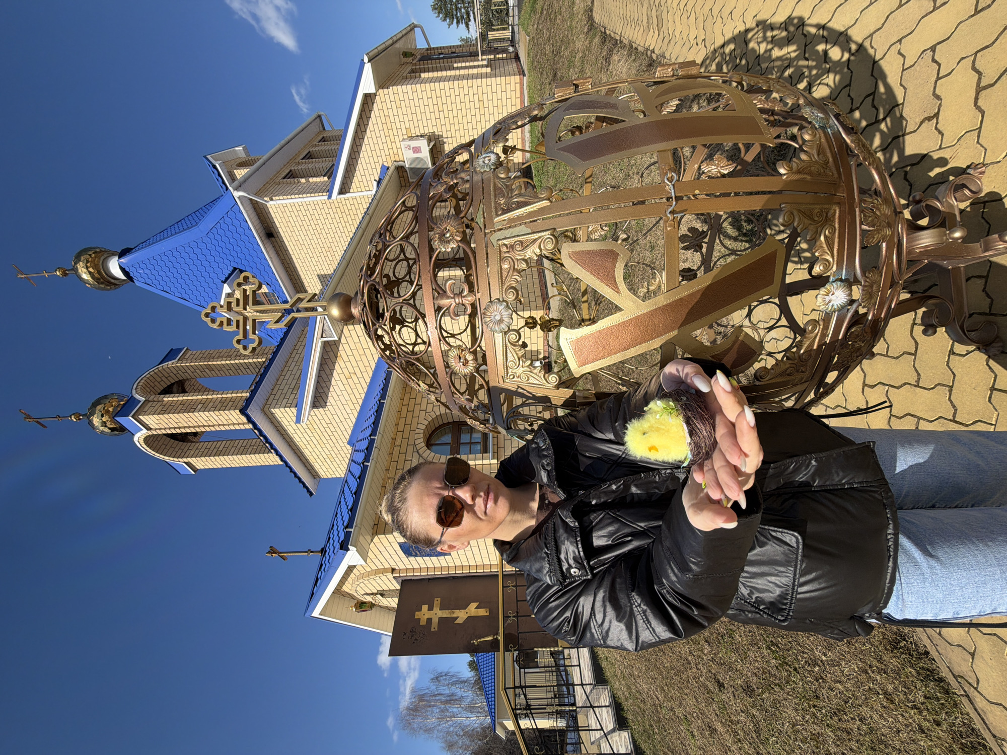 The image shows a woman in a black jacket and sunglasses standing outside the Church of the Holy New Martyrs and Confessors of Russia in Chaikovsky, Russia. She is holding a small object with a yellow chick in her hands. Behind her is a large, ornate golden structure with Cyrillic characters. The church is constructed of light-colored brick with blue roofing and golden domes topped with crosses. The sky is blue with some clouds, and the ground appears to be paved with stone tiles.