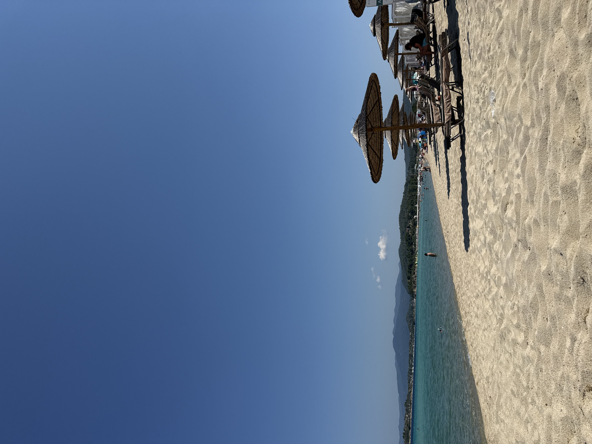 This is a photograph of a sandy beach on a clear, sunny day. The scene is dominated by the bright blue sky on the left, contrasting with the sandy beach and turquoise water on the right. A row of straw-thatched umbrellas and lounge chairs are set up on the sand, casting long shadows, indicating it is likely mid-day. Several people are visible on the beach, some relaxing under the umbrellas, and others in the shallow water. In the background, a line of green, tree-covered hills or mountains stretches along the coastline. The water is calm and inviting, with a few small waves lapping at the shore. The overall atmosphere is one of leisure and relaxation, typical of a summer beach day in a Mediterranean location like Sithonia, Greece. There are no visible signs or text in the image.