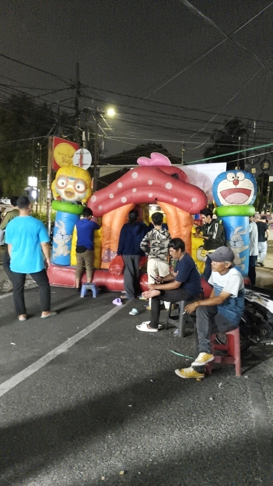 This image captures a nighttime street scene in South Jakarta, Indonesia, likely during a local event or market. The central focus is a large, inflatable bouncy castle with characters resembling Pororo and Doraemon. Several people, mostly young men, are present. One man in a blue t-shirt stands facing away from the camera, while others are gathered around or near the bouncy castle. Some appear to be supervising or waiting, while others are interacting with their phones. One man is seated on a red stool in the foreground, also looking away from the camera. The environment suggests a temporary setup on a paved road, with streetlights and overhead wires visible, indicating an urban setting. There's a sign with red and yellow elements to the left, though its text is mostly illegible. The overall atmosphere appears casual and community-oriented, common in South Jakarta's vibrant night markets and local gatherings.