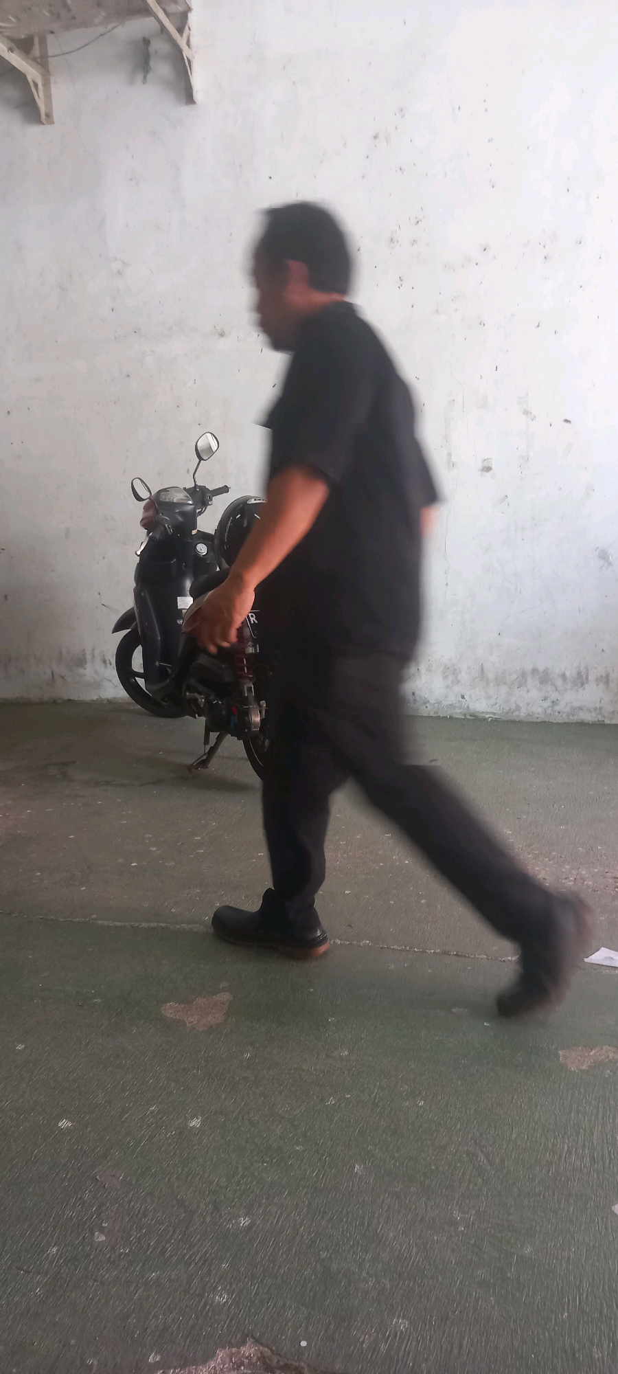 A person wearing a black shirt and black pants is walking past a parked black motorcycle. The person's face is blurred due to motion. The motorcycle has a silver mirror and a black helmet on the seat. The scene is set against a textured, off-white wall and a green, worn floor. The image appears to be taken in an indoor or covered outdoor space, possibly a garage or parking area. The presence of a motorcycle and a stark wall suggests a utilitarian environment. The specific location in Banjarnegara, Indonesia, is not discernible from the visual elements alone; however, the overall scene depicts an everyday moment of transit or activity.