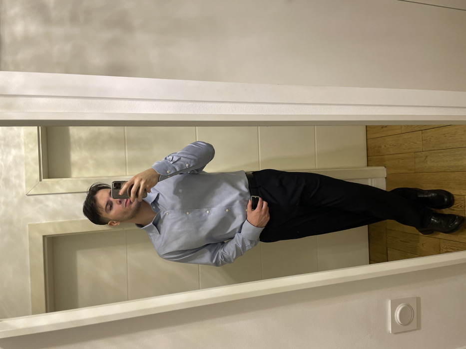 A man in a light blue dress shirt and dark trousers stands in front of a mirror, taking a selfie with his phone. He has dark hair and a serious expression. The background includes a tiled wall, a wooden floor, and a light switch on the wall. The mirror frame is white.