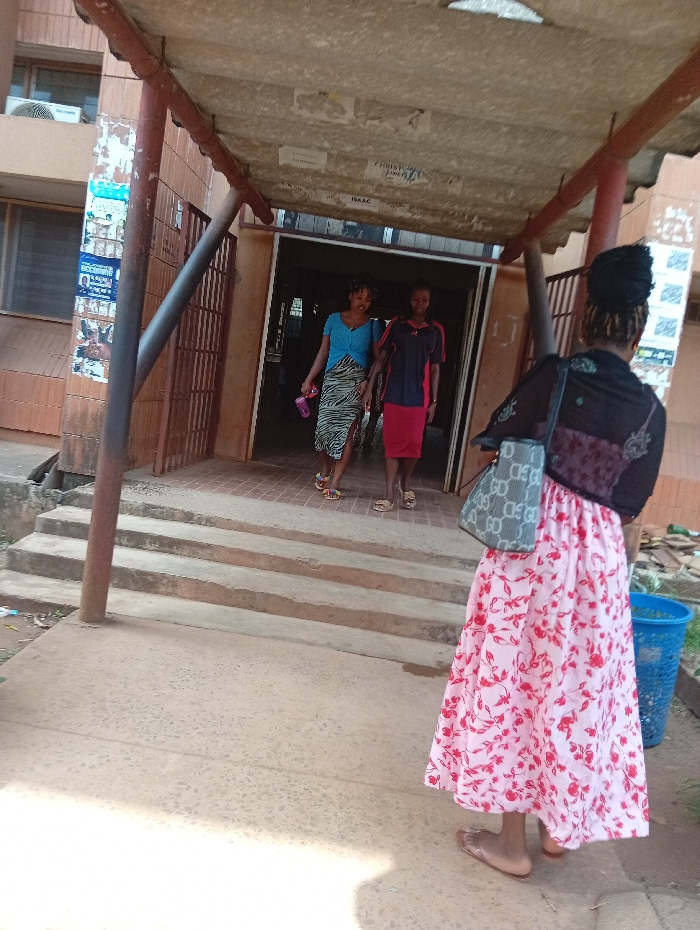A daylight outdoor scene in Nsukka, Nigeria, depicts three individuals near a building entrance with concrete steps and an overhead canopy. In the foreground, one individual, seen from behind, wears a black top, a long pink skirt with red floral patterns, and light-colored sandals, carrying a patterned shoulder bag on her left side. She faces a building entrance. From this entrance, two individuals walk down three concrete steps towards the foreground. The person on the left wears a light blue top, a patterned skirt, and light-colored sandals, holding a pink bottle. The person on the right wears a dark polo shirt with red accents, a red skirt, and light-colored sandals. The building's left wall features brown tiling, a window, an air conditioning unit, and various posters. The underside of the overhead canopy is discolored and has posters affixed, showing partial text like 'CHRIST' and 'ISAAC'. A blue plastic bin is visible to the right of the steps. The ground is a light-colored concrete slab.