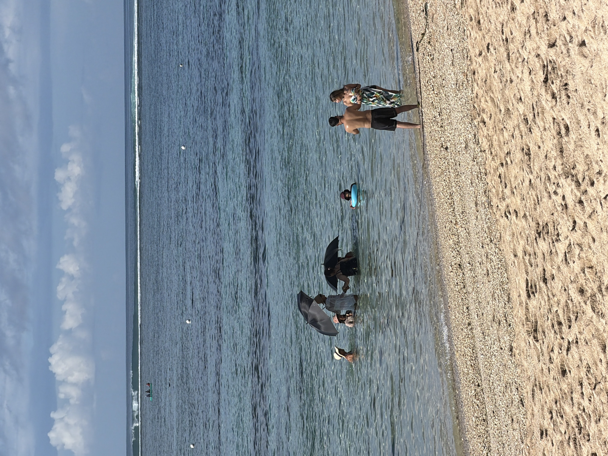The image depicts a coastal scene in Unknown, Indonesia, showing a beach with a mix of sand and small pebbles transitioning into clear water. In the water, several people are present. Some are partially submerged, holding black umbrellas, while another person is wearing a hat. Further out in the water, a person is in a blue inflatable ring. Near the shoreline, a shirtless man in dark shorts stands next to a woman in a floral dress. The water extends out towards a horizon line visible in the distance. The sky is partially cloudy with white clouds visible. There are distant objects in the water, possibly buoys. Overall, the scene appears to show people enjoying a sunny day at the beach.