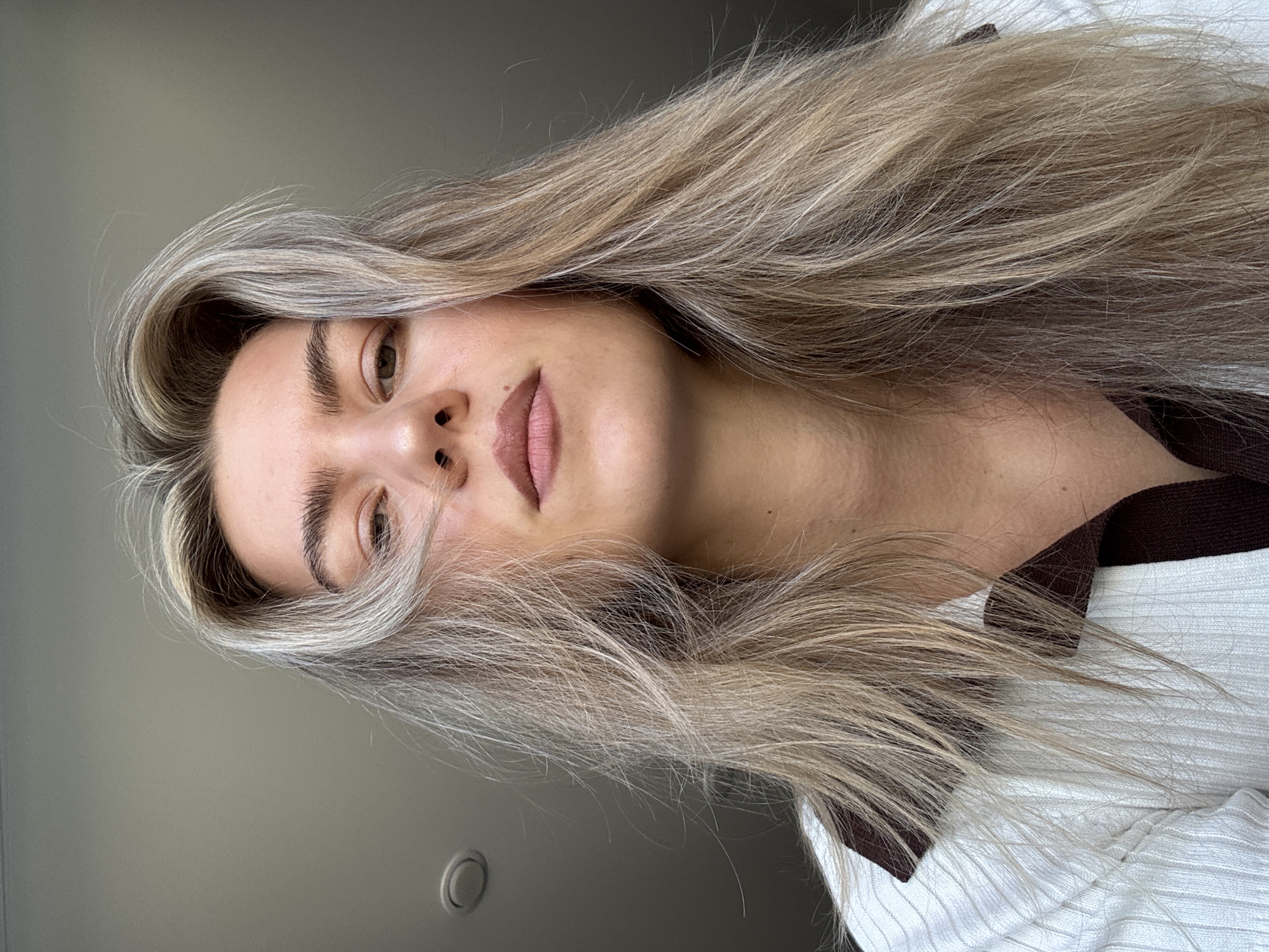 A woman with blonde hair is facing the camera. She has light brown eyes, a small nose, and pink lips. She is wearing a white ribbed shirt with a brown collar. The background is a gray wall with a circular object on it. The location is unknown.