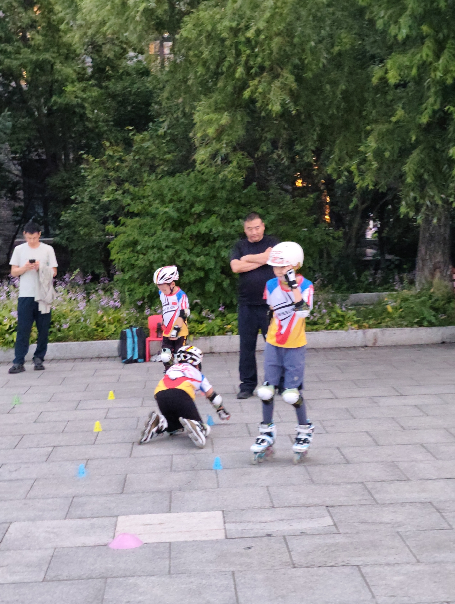 In the city of Aihui, China, three children are practicing rollerblading on a paved plaza. Two of the children are wearing full protective gear, including helmets and knee pads. One child is on their knees, while the other two are standing. A man stands in the background, observing the children. The area is surrounded by trees and some flowering bushes, suggesting an outdoor park or recreational area. The plaza is paved with large, rectangular stone tiles. Several colored cones are scattered on the ground, indicating a training course. A backpack and a red chair are visible near the rollerblading area. The scene appears to be during the daytime, with soft lighting.