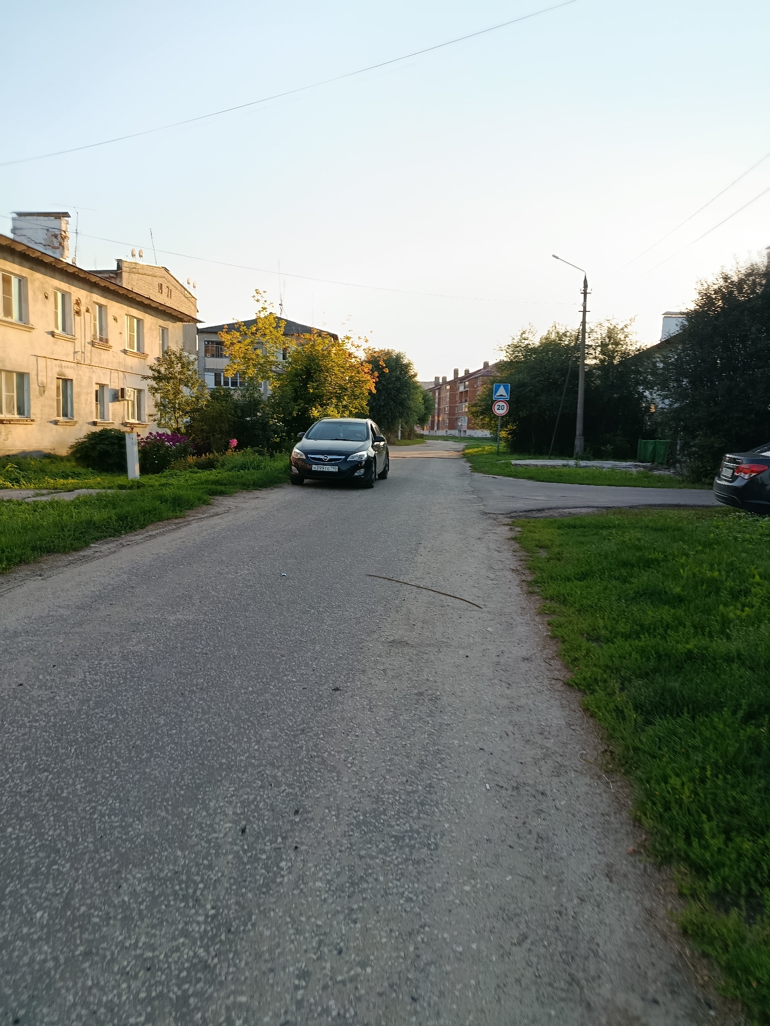 The image shows a street scene in Vareya, Russia, likely during daylight hours with clear skies. In the foreground, a grey asphalt road occupies the majority of the frame, with a strip of green grass lining the right side. Several apartment buildings are visible on the left side of the road, appearing to be of Soviet-era architecture. A dark-colored passenger car is parked on the side of the road in the mid-ground, with its front facing towards the camera. Further down the road, a traffic sign indicating a speed limit of 20 is visible. Trees and bushes are scattered throughout the scene, some with autumn foliage, suggesting it might be late summer or early fall. The overall environment appears to be a residential area within a town or small city.