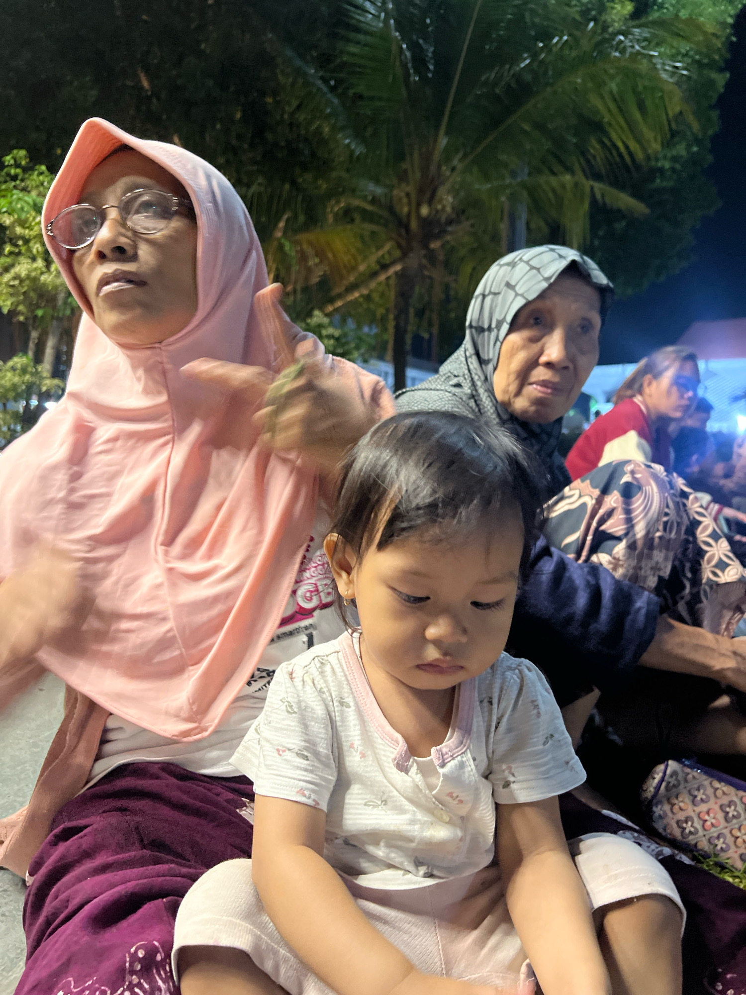 In the city of Kecamatan Pati, Indonesia, at night, a group of people are gathered outdoors. In the foreground, a young child with dark hair and light skin sits facing forward, wearing a white t-shirt with a pink collar and a small pink hair accessory. To the left and slightly behind the child, an adult woman wearing a pink hijab and glasses looks upwards and to the left, her right hand raised. Behind her and to the right, an elderly woman in a grey and white patterned hijab looks towards the left. Further in the background, another woman in a red and white top is visible, along with other figures in the dim light. The setting appears to be a public space with trees, possibly a park or a street, illuminated by artificial lights. The ground is covered with what appears to be a dark purple cloth or blanket.