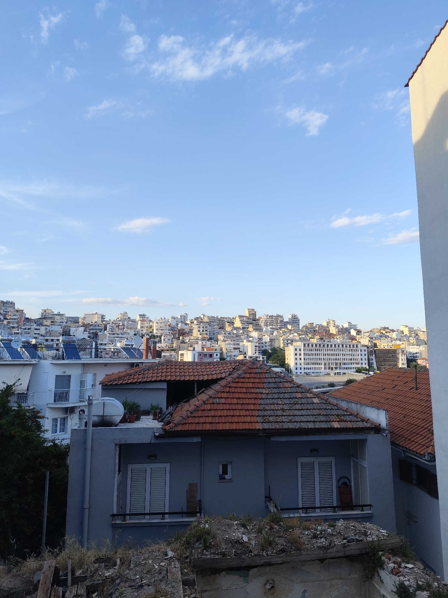 The image captures a cityscape with a clear blue sky dotted with sparse white clouds. In the foreground, a gray house with a red-tiled roof is visible, featuring windows with closed white shutters and a small balcony. To the left, another white building with balconies and windows is partially visible, adjacent to lush green trees. Behind these structures, a dense collection of multi-story buildings, typical of an urban environment, stretches across the horizon. The architecture appears to be a mix of older and modern styles, with many buildings featuring balconies and varying roof types. The presence of satellite dishes on some rooftops indicates a residential area. The scene suggests a daytime view in an urban setting, likely Greece given the architectural style and the context of Kavala. The foreground also shows remnants of construction or demolition with debris scattered on a lower level.