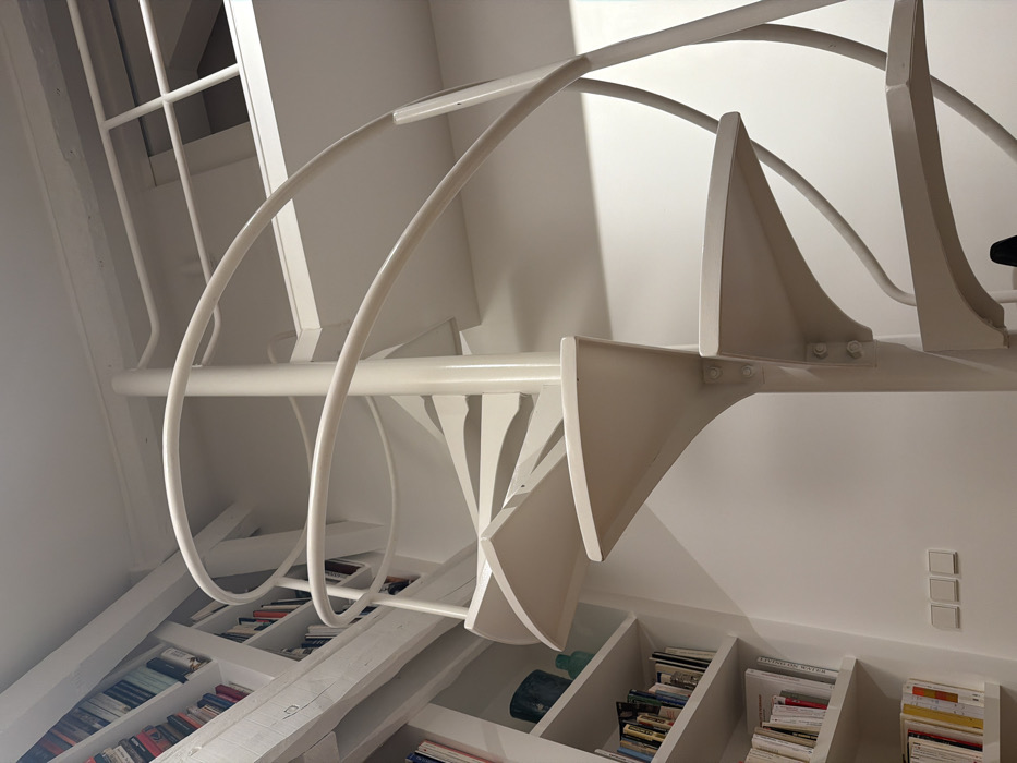 This image depicts a brightly lit interior space, likely within an apartment or studio in Paris, France. The central subject is a cream-colored metal spiral staircase with a central pole and curved tubular handrails. The individual steps are solid, triangular in shape, and appear to be bolted to the central support structure.
To the left of the staircase, a floor-to-ceiling white bookshelf system is integrated into the wall, filled with numerous books of various sizes and colors. On one of the upper shelves, two decorative vases, one teal and one lighter blue-green, are visible. Exposed, white-painted wooden beams run horizontally along the top left portion of the ceiling, indicating structural elements. Below the bookshelves, on a white wall, three rectangular white light switches are installed horizontally.
Above and to the right of the staircase, a mezzanine or loft area is visible, defined by a white railing system with vertical and horizontal bars. The walls and ceilings throughout the space are predominantly white, contributing to a clean, modern aesthetic. There are no individuals, animals, or discernible actions or interactions occurring within the frame.