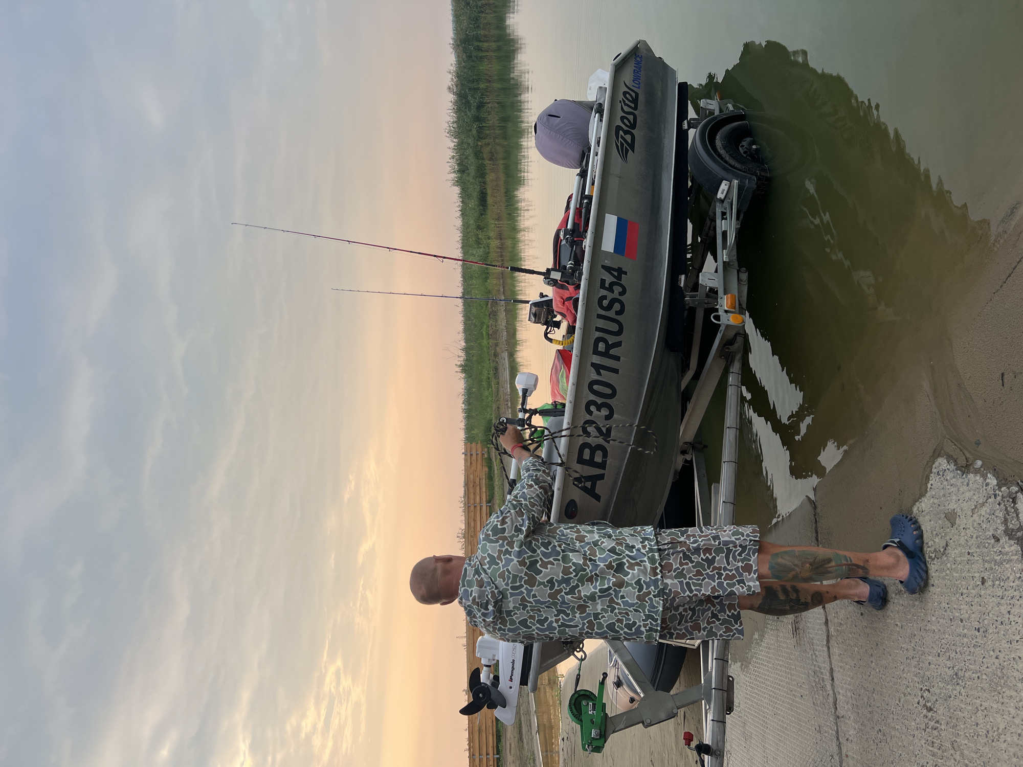 A bald man in camouflage clothing stands next to a boat on a trailer at the edge of a body of water. The boat is silver with the license plate "AB2301RUS54" and a Russian flag sticker on its side. Fishing rods are placed in the boat, and a trolling motor is attached to the stern. The man appears to be preparing the boat, holding ropes in his hands. The environment consists of a sandy shore, concrete ramp, and lush green vegetation in the background. The sky is overcast with hints of pink and orange from a sunset. The trailer's wheels are partially submerged in the water, suggesting the boat is being launched or retrieved. The image appears to be taken in Zav'yalovo, Russia, given the license plate.