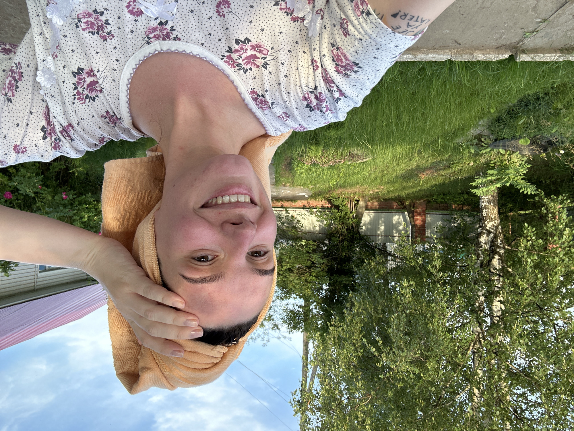 A young woman with a tan towel wrapped around her head poses for a selfie outdoors. She is wearing a white t-shirt with a floral pattern. Her left arm is raised, touching her forehead. The background features a grassy yard, a tree, a fence, and a house with a pink roof. The scene appears to be in a rural or suburban area, possibly in Альняш, Russia, given the architectural style and vegetation. The lighting suggests it is daytime, likely late afternoon due to the warm glow. A tattoo is visible on her left forearm, with the text "SEE YA LATER!"