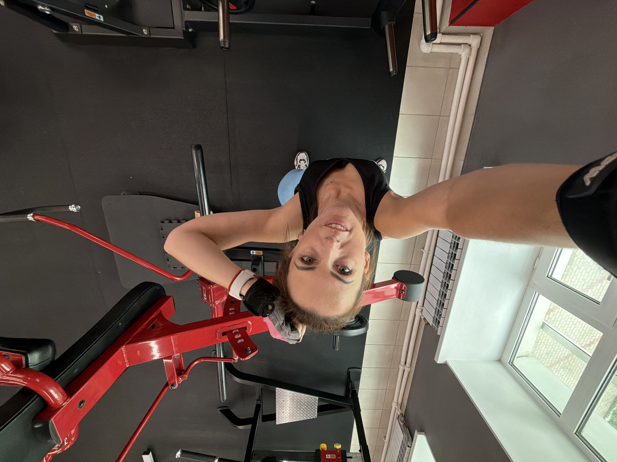 An inverted, first-person perspective selfie taken in a gym. A woman is looking directly at the camera, smiling. She is wearing a black tank top, light blue athletic leggings, and white and black athletic shoes. She has on black fingerless gloves with a white band on her left wrist. Behind her, a red weight machine is visible, along with black gym flooring and a section of tiled wall. In the foreground, the arm of the person holding the camera is visible. The scene appears to be a gym in Birobidzhan, Russia.