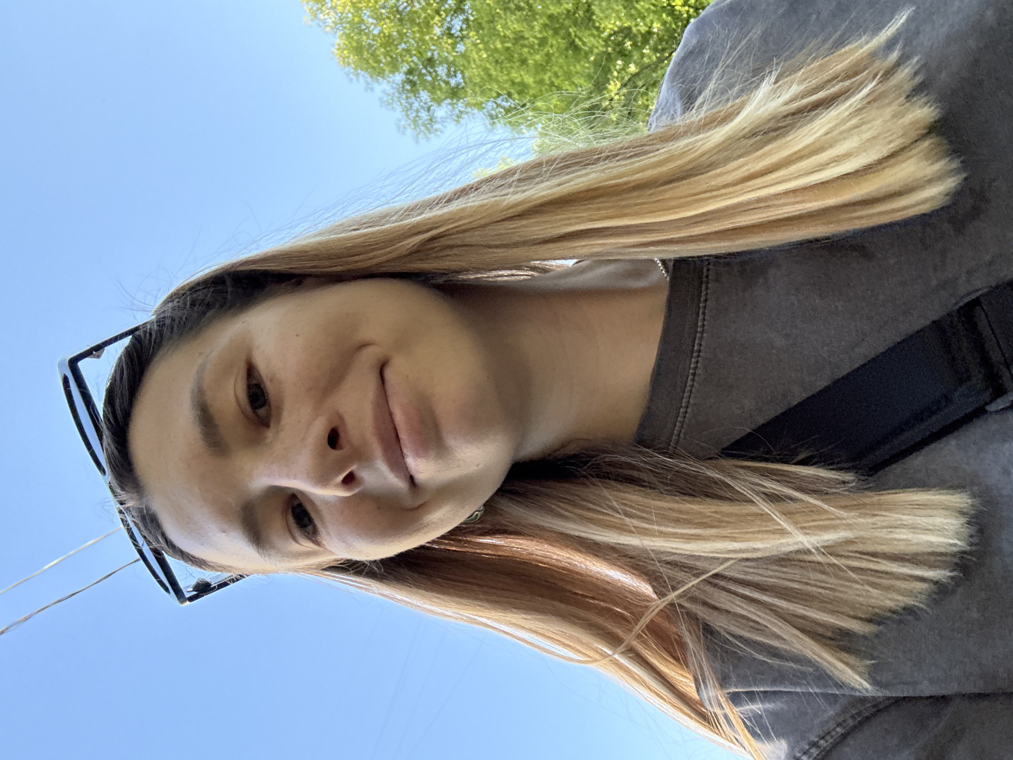 A young adult female is depicted in a low-angle outdoor selfie, looking directly at the camera with a subtle smile. She has long, straight, blonde and light brown hair with highlights, extending past her shoulders. Black-framed sunglasses are pushed up onto her head. Her face displays clear skin with visible freckles. She wears a dark grey, crew-neck t-shirt or sweatshirt with a black strap, possibly from a bag, diagonally across her chest. A thin, silver-colored chain necklace and a small metallic earring in her left ear are visible. The background features a clear, bright blue sky with two thin, parallel wires visible in the upper left. Lush green tree foliage is present in the upper right. The scene is set in Moscow, Russia.