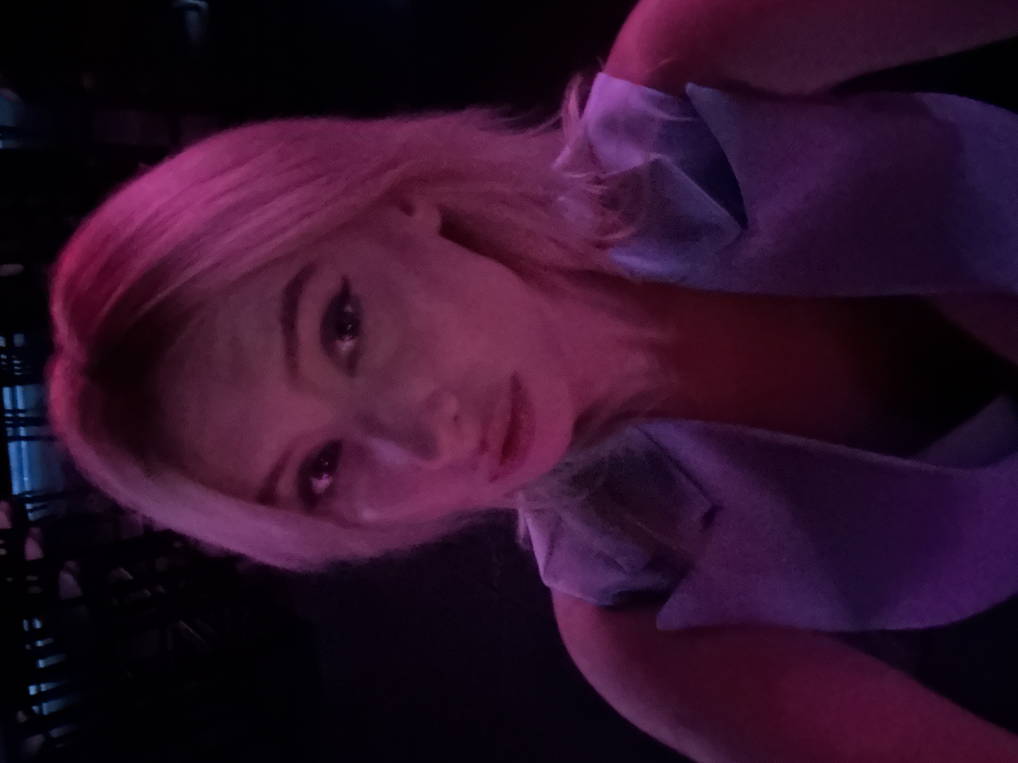 The image is a close-up, selfie-style photograph of a young woman with blonde hair, illuminated by pink and purple lighting, suggesting a dimly lit indoor environment. She is wearing a light blue vest. The background is dark and indistinct, with hints of blurry shapes that could be furniture or other patrons, indicating a social setting like a bar or club. There is no visible indication of the city of Amman, Jordan, or any specific geographic markers.