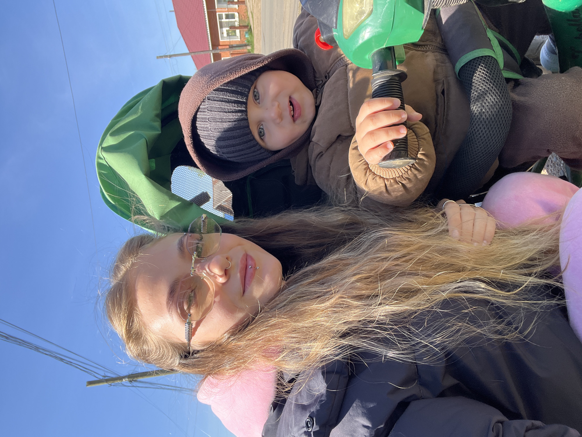 A mother and child are posing for a photograph outdoors in what appears to be a residential area. The mother, wearing glasses and a dark jacket with a pink accent, is positioned with her face toward the camera. Her long, blonde hair is spread out around her shoulders. The child, wearing a brown hooded jacket and a gray knit hat, is holding onto part of a green stroller or child carrier. A house with a red roof is visible in the background. Above the figures is a bright blue sky. The location appears to be in the city of Фоки, Russia.