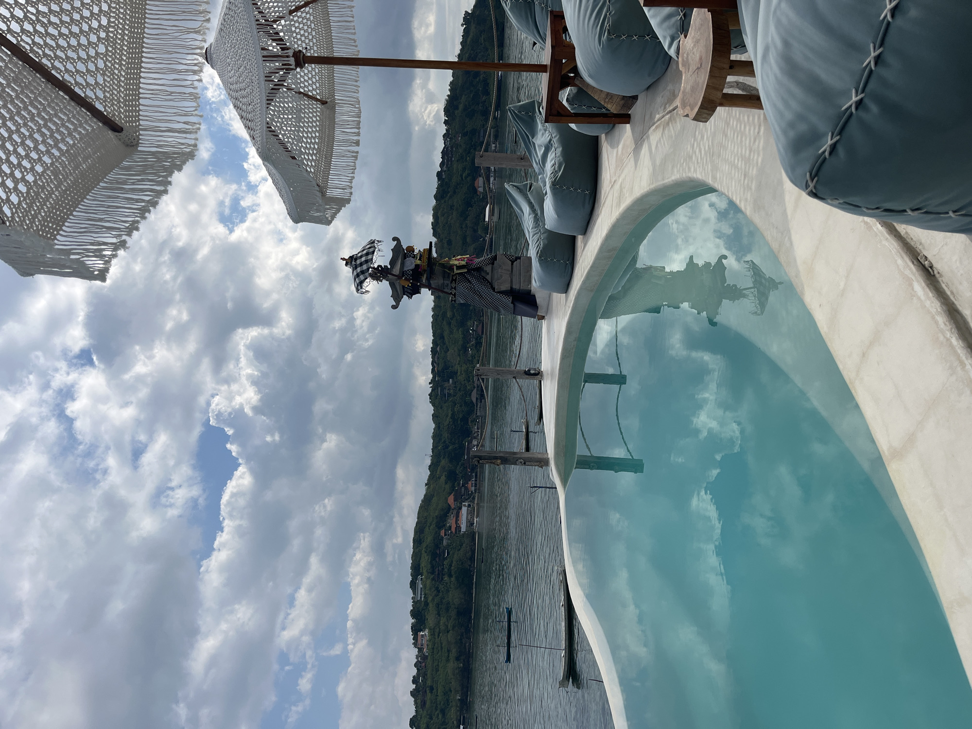 This is a medium shot, taken from a slightly high angle, of an infinity pool overlooking a body of water. The pool is in the foreground, its turquoise water reflecting the cloudy sky. To the right of the pool, on a white concrete deck, are several light blue beanbag chairs and a small wooden table. In the background, across the water, is a green, hilly coastline with scattered buildings. Several small boats are visible on the water. Two white umbrellas with decorative fringe are partially visible at the top of the frame, suggesting an outdoor seating area or resort. The scene appears to be in Nusa Penida, Indonesia, given the distinctive landscape and the Balinese-style altar-like structure visible behind the pool on the left.