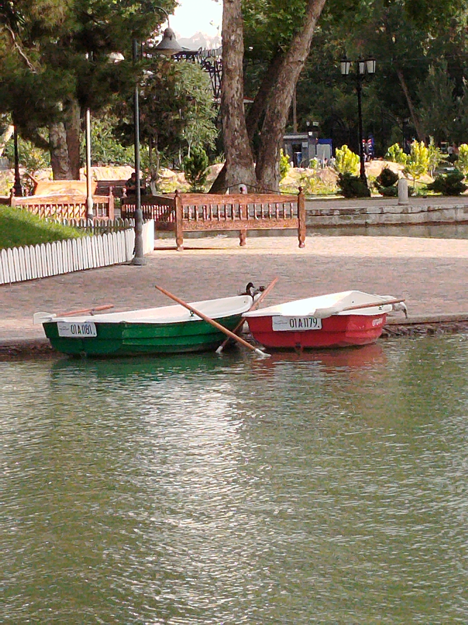 Two rowboats are docked on the edge of a body of water in a park. The boat on the left is green with white interior and is labeled "01A 1181". The boat on the right is red with a white interior and is labeled "01A 1179". A wooden bench is visible behind the boats, along with a white picket fence. In the background, there are trees and lampposts, and a building. This scene is located in Tashkent, Uzbekistan, likely in Amir Timur Square, which features a large pond with rowboats.