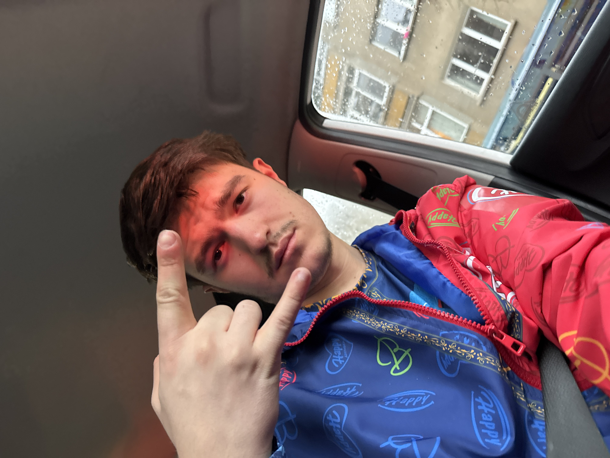 A young adult male is taking a selfie in a vehicle. The photo is taken from a low angle, looking up at the subject. The subject is wearing a blue jacket with "Happy" written on it and a red jacket with colorful logos on top. He is making a hand gesture, holding up his index and pinky fingers. Outside the vehicle, through a rain-streaked window, a multi-story building with multiple windows can be seen. Based on the architecture and presence of Cyrillic script on building signs, the location is consistent with Sofia, Bulgaria. Rain is visible on the window.