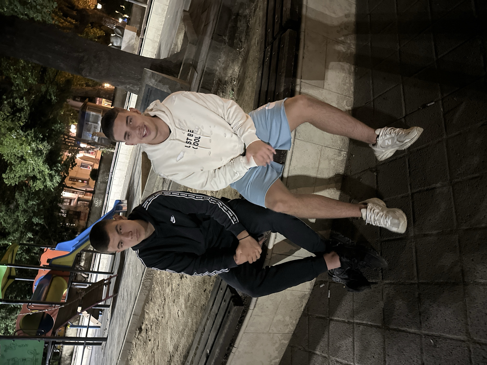 Two young men are positioned diagonally across the frame in an outdoor setting in Pazardzhik, Bulgaria. The man on the top is wearing a white hoodie with the print "UST BE (100)", light blue shorts, and white sneakers. The man below him is wearing a black Nike tracksuit and black sneakers. A playground slide is visible in the background on the left side, and trees and buildings are present in the distance. The ground appears to be paved with dark-colored tiles. The lighting suggests it is nighttime.