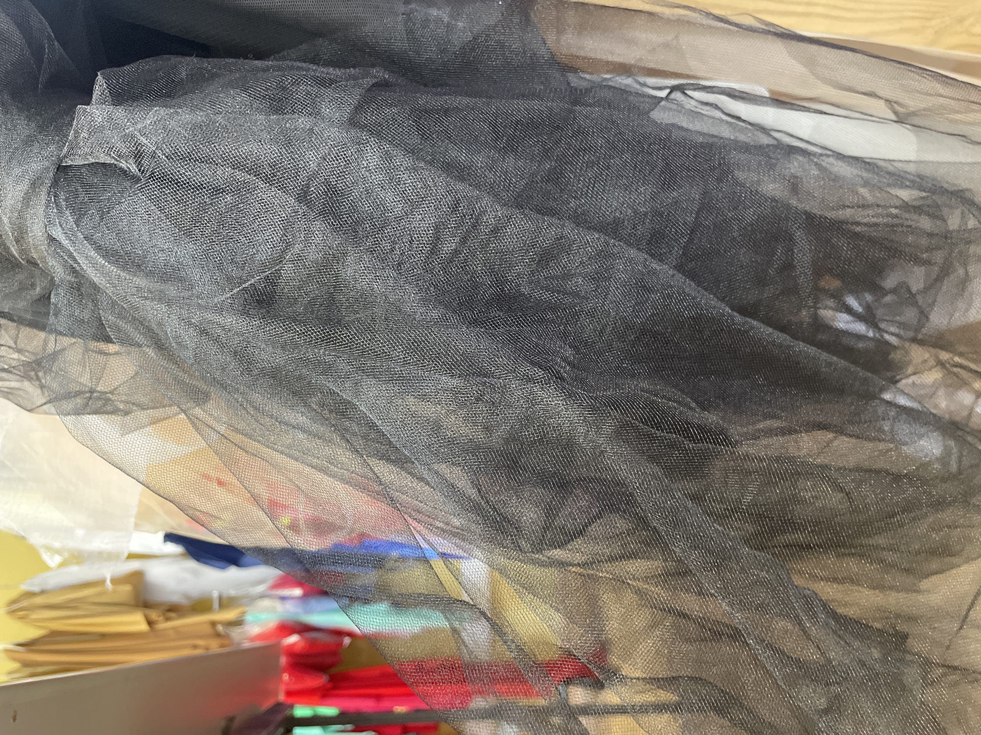 The image shows a close-up of black tulle fabric. The fabric is layered and has a mesh-like texture. In the blurred background, several colorful garments are visible hanging on a rack, including red, blue, and gold clothing. The context provided about Sinyavino, Russia is not evident from the visual information in the image.