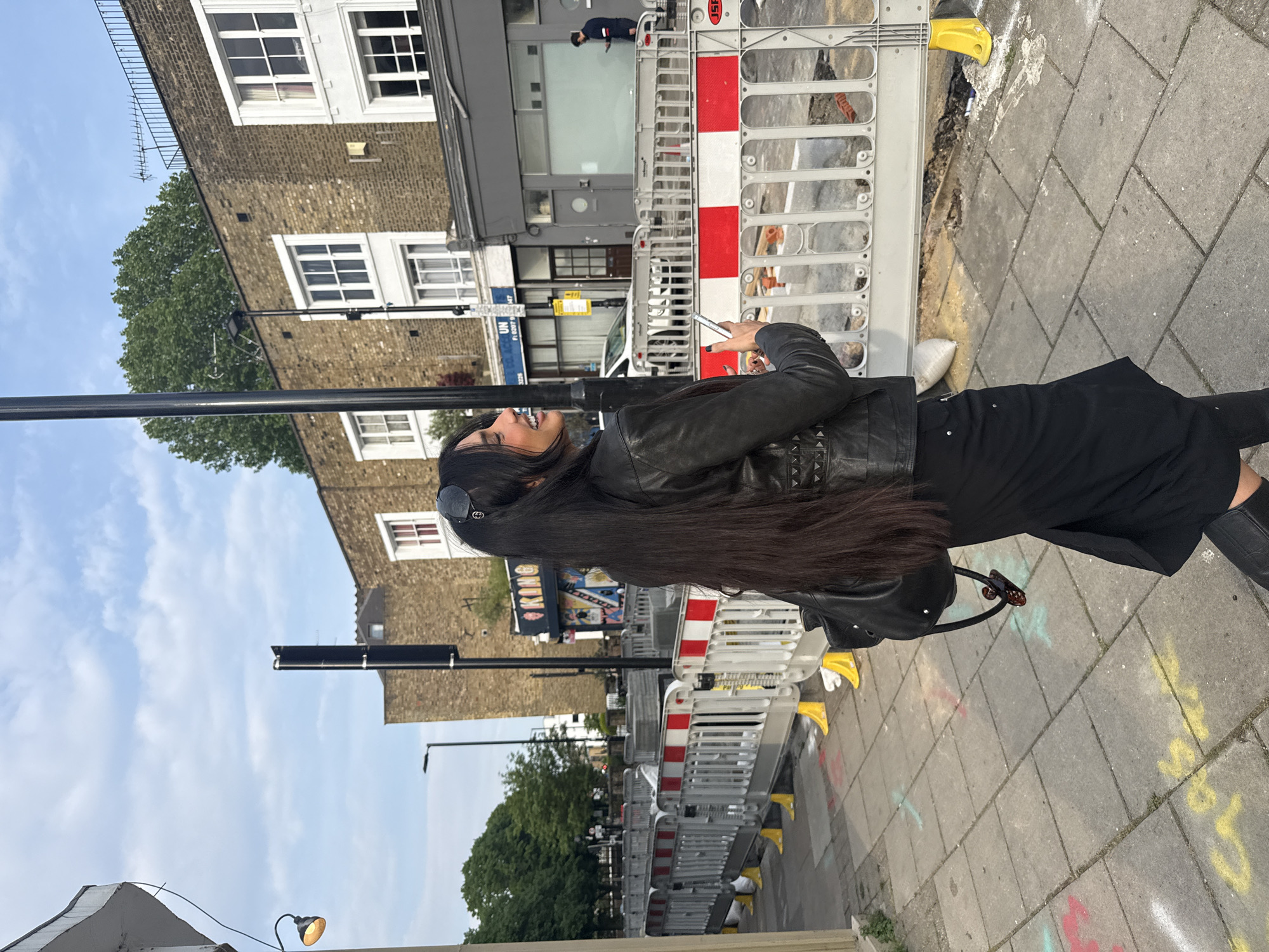 A woman with long, dark hair is standing on a paved sidewalk in what appears to be a street in London, United Kingdom. She's wearing a black leather jacket and a black skirt or dress. She is smiling and holding a mobile phone. Road work barriers are visible along the sidewalk, painted with red and white stripes and yellow supports. Buildings of brick and light-colored stone form the backdrop. The sky is pale blue with some clouds. Street signs and other details characteristic of a city environment, like lampposts, are present.